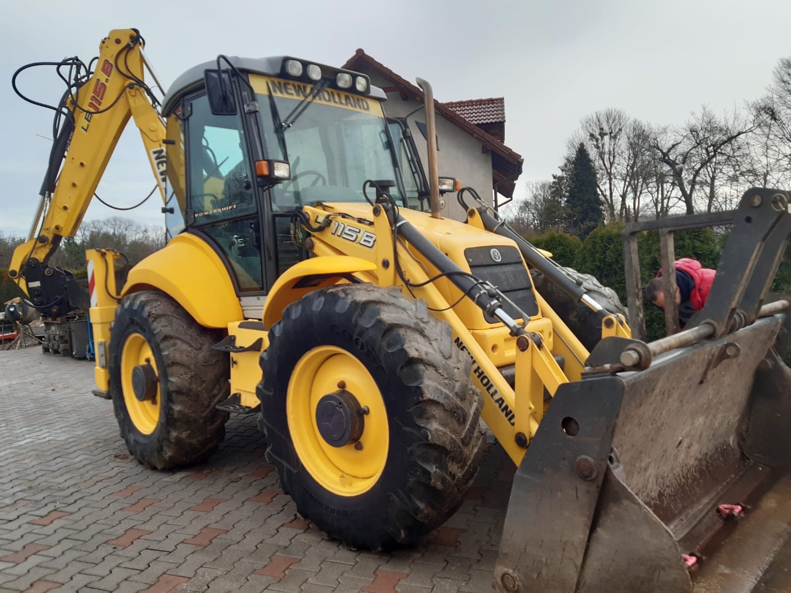 Żółta koparko-ładowarka New Holland 115B na kostce brukowej, z widocznym operatorem w kabinie i elementami otoczenia, takimi jak budynek i drzewa.