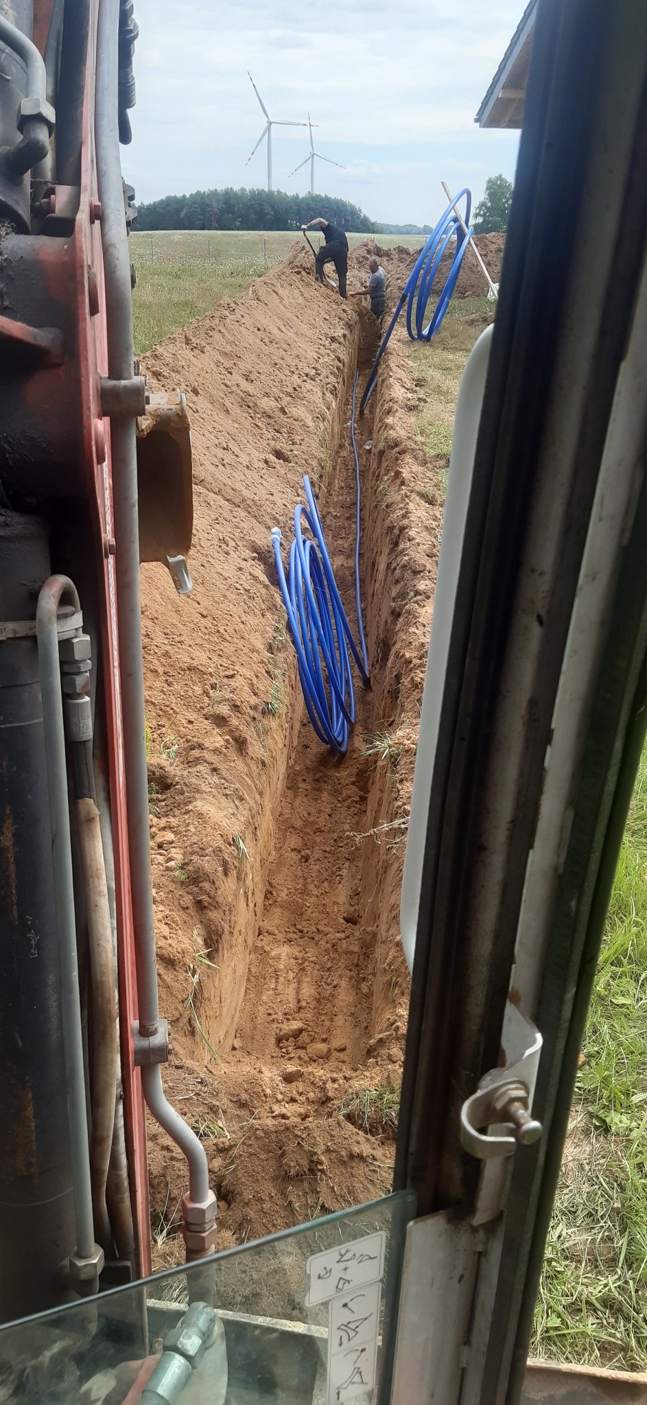 Wykopany rów z niebieskimi rurami w ziemi, widok z kabiny koparki. W tle pracownicy i turbiny wiatrowe. Prace ziemne w toku.