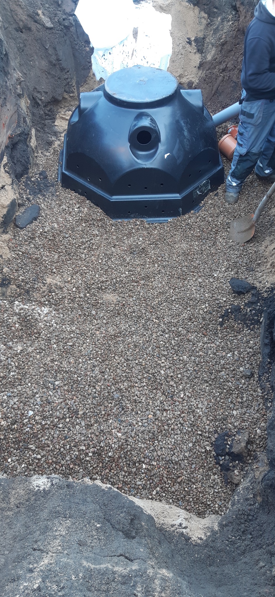 Montaż czarnego zbiornika na szambo betonowe w wykopie, podsypka żwirowa, widoczne rury i fragment pracownika. Realizacja przyłącza kanalizacyjnego.