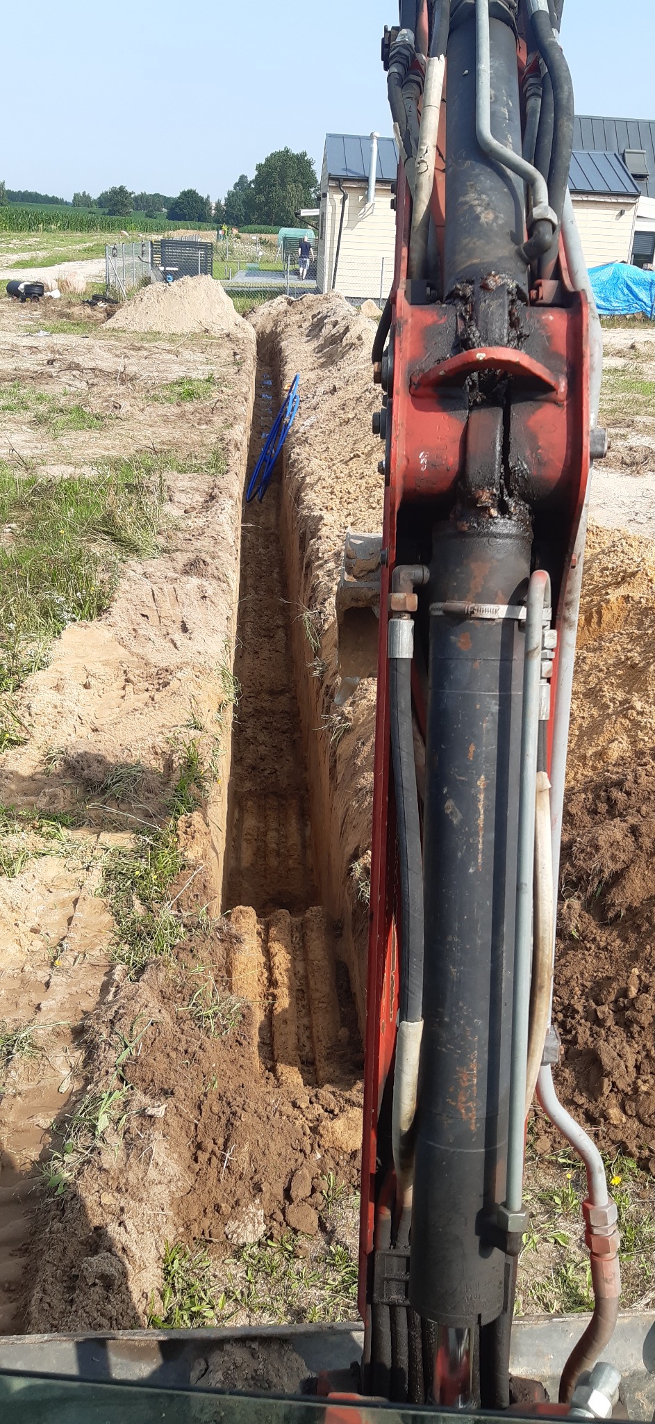 Wykopany wąski rów w ziemi, z widocznymi niebieskimi rurami na dnie, wykonany prawdopodobnie koparką, której ramię jest widoczne na pierwszym planie.