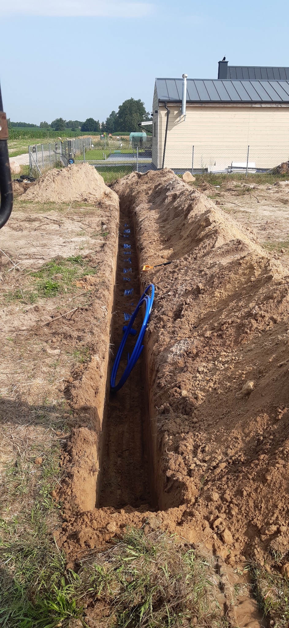 Wykopany rów z ułożonymi niebieskimi rurami instalacyjnymi na terenie budowy, widok z góry. W tle budynek mieszkalny i pole uprawne.