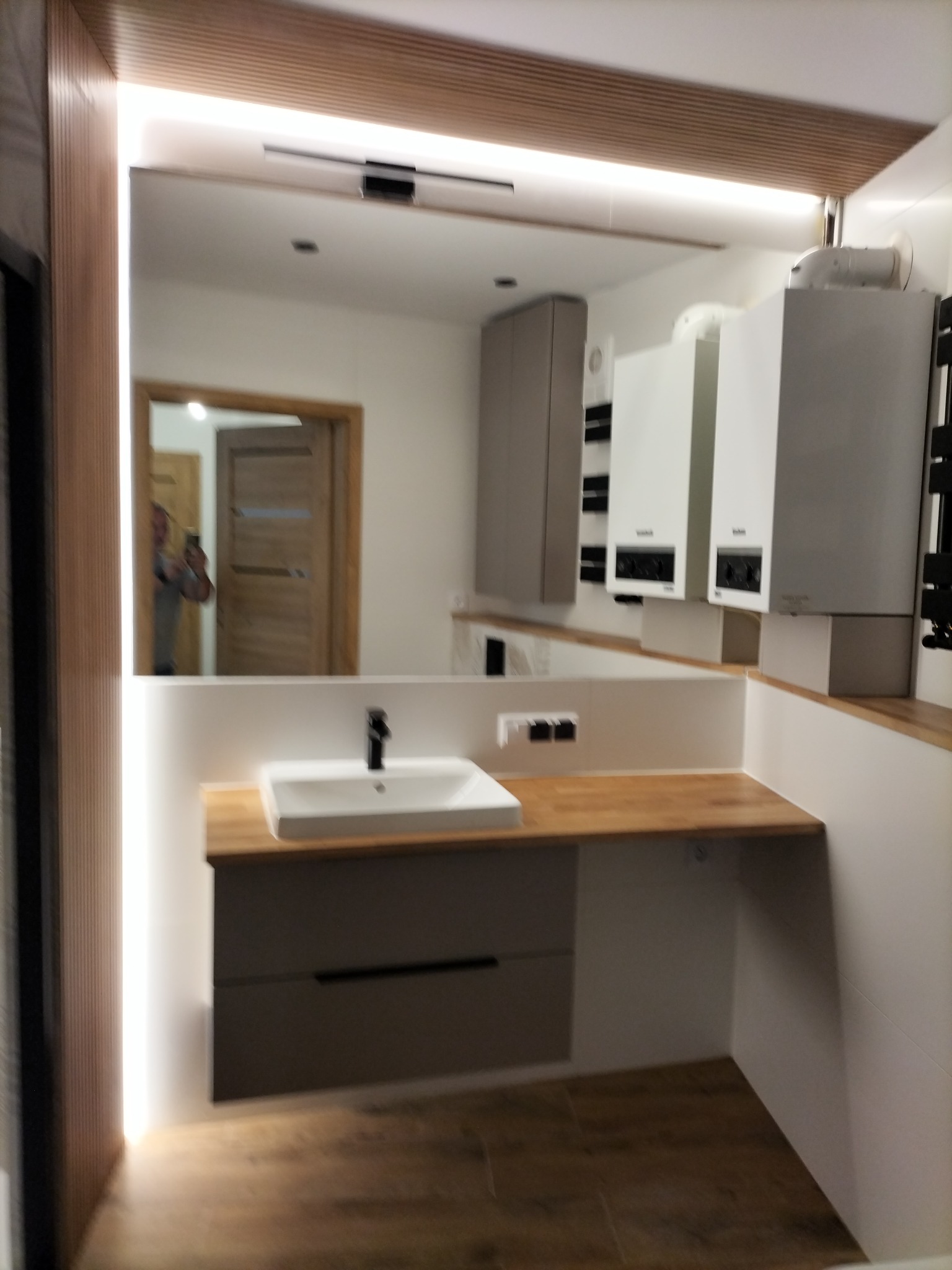 Nowoczesna łazienka z podświetlanym lustrem, drewnianym blatem i szarą szafką pod umywalką. Widoczne bojlery i drzwi w lustrzanym odbiciu. Minimalistyczny design.