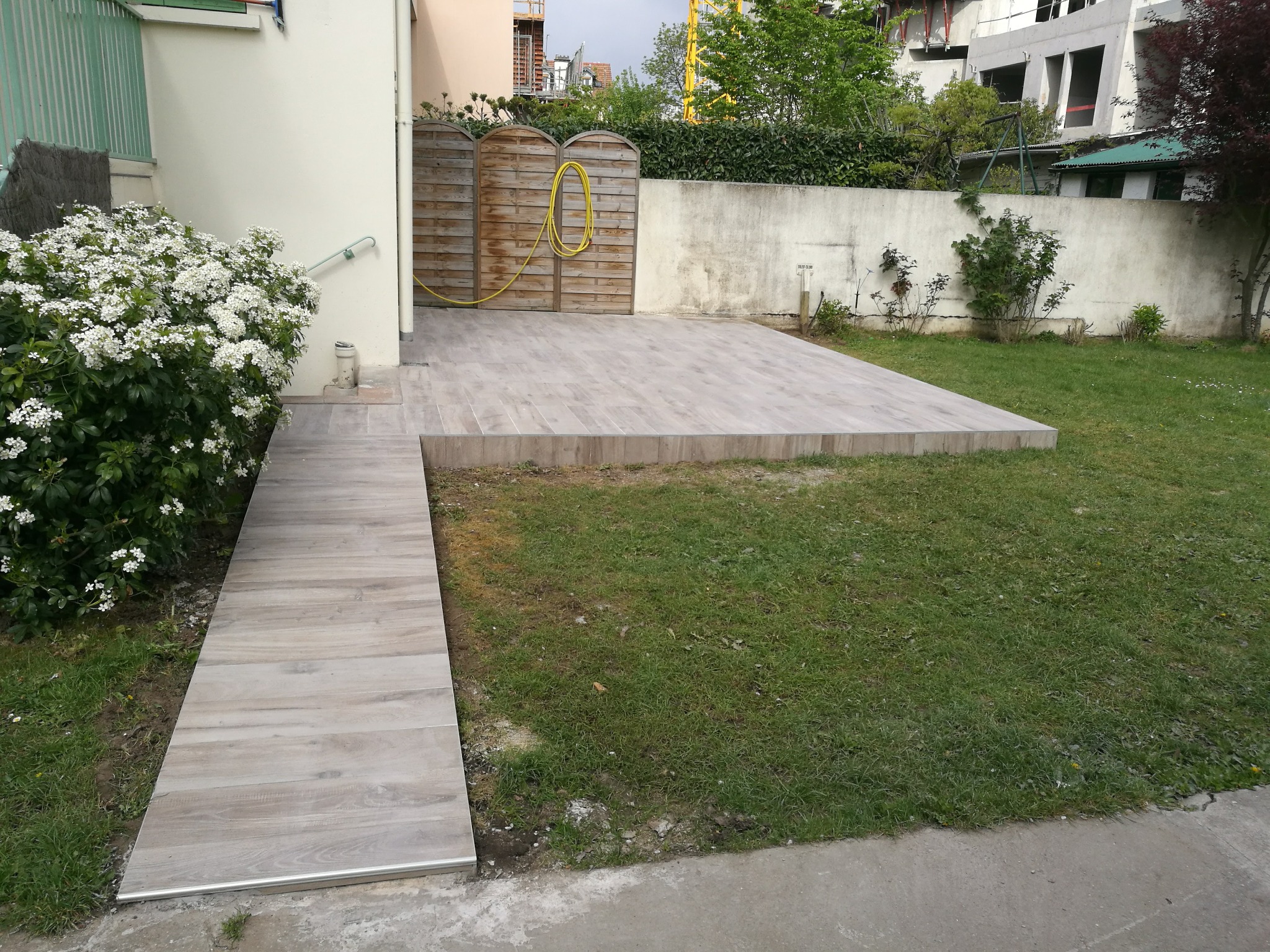 Wykończony taras z płytkami imitującymi drewno i rampą w Stalowej Woli. Widoczny ogród i ogrodzenie. Żółty wąż ogrodowy zwisa z drewnianej bramy.