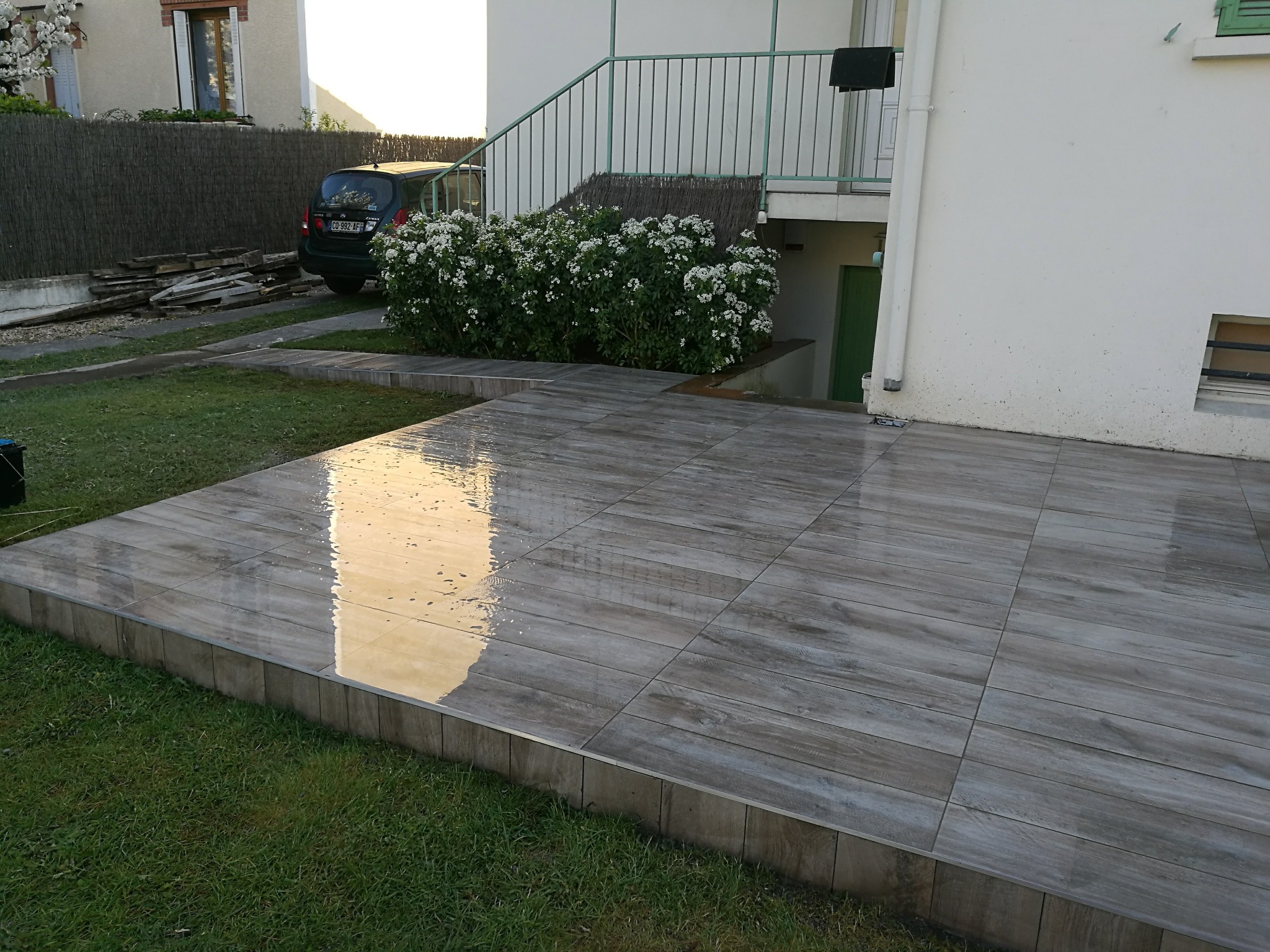 Nowoczesny taras z szarych płytek imitujących drewno, z widocznym odbiciem światła, otoczony zielonym trawnikiem i roślinnością. Schody prowadzą do budynku.