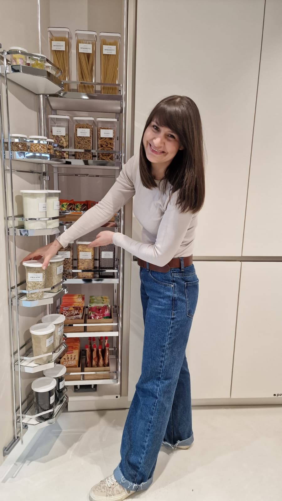 Uśmiechnięta kobieta prezentuje wysuwaną spiżarnię z zapasami żywności w pojemnikach. Porządek i organizacja w kuchni.