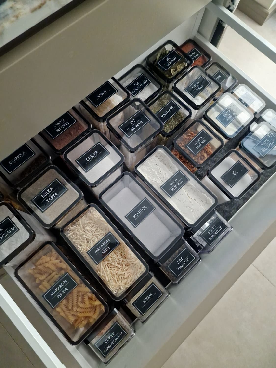 Szczelnie zamknięte, przezroczyste pojemniki z etykietami w szufladzie kuchennej. Uporządkowane przyprawy i produkty sypkie, estetyczne przechowywanie.