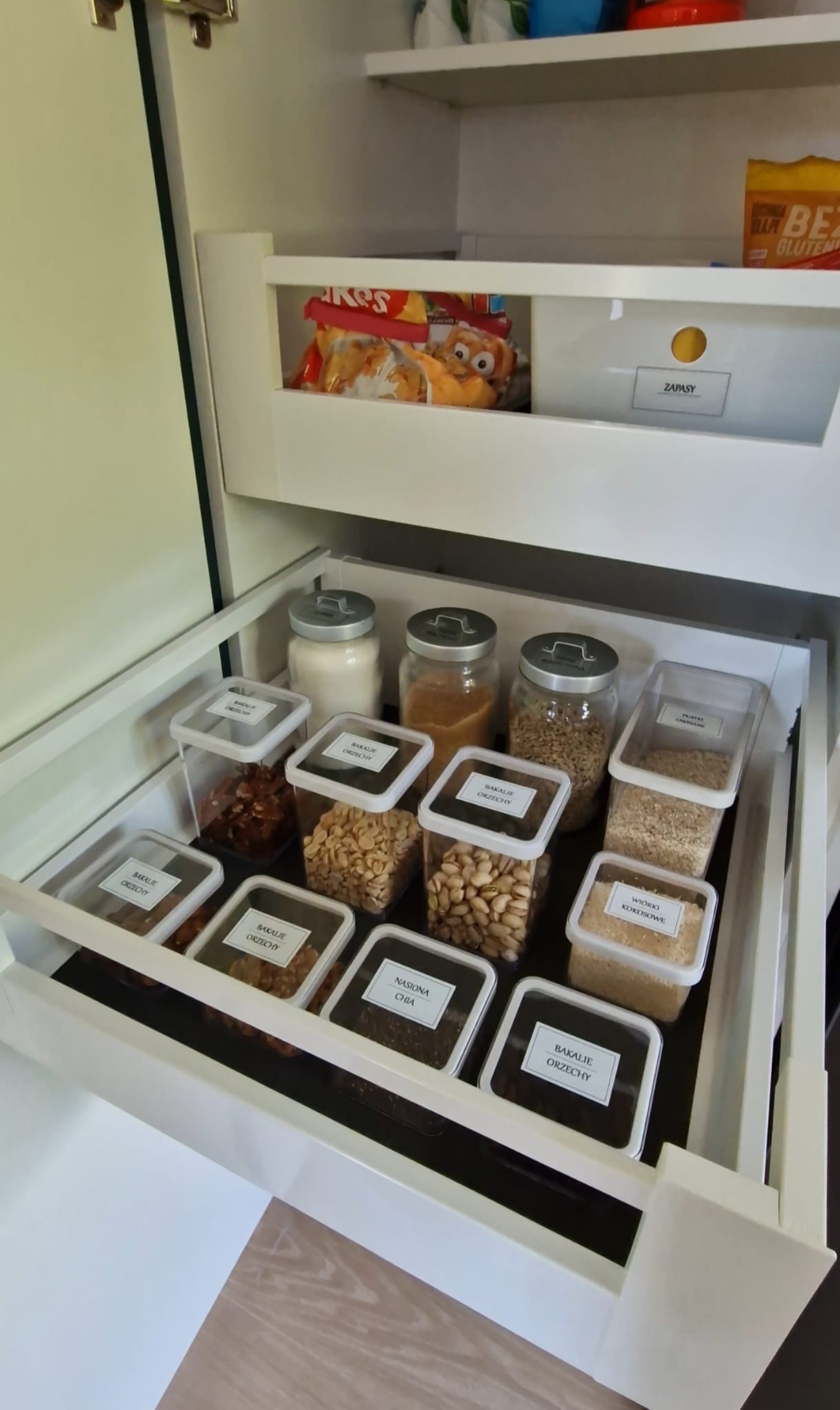 Półka zorganizowana w szafce kuchennej. Przezroczyste pojemniki z etykietami na bakalie i orzechy. Porządek w zapasach.