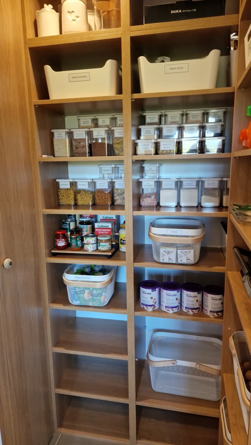 Spiżarnia z drewnianymi półkami, na których poukładane są pojemniki z suchymi produktami, konserwami i koszyki. Organizacja przestrzeni w kuchni.