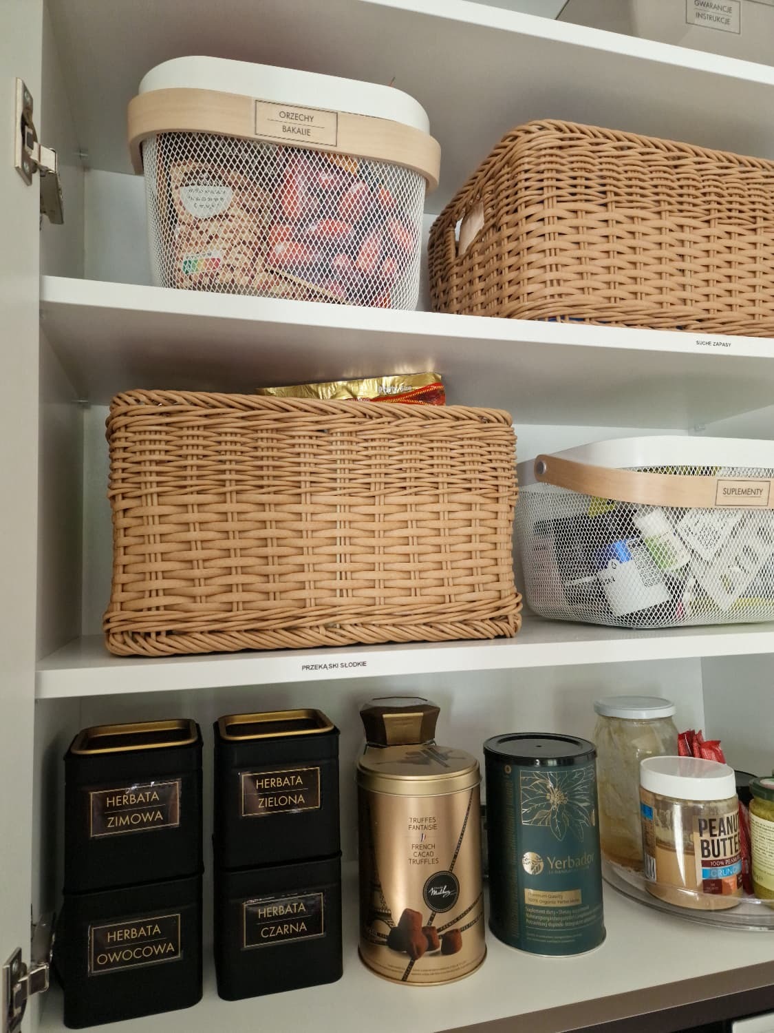 Półka w szafce spiżarni po uporządkowaniu: koszyki z orzechami, suplementami, pudełka herbat i słoiki z przekąskami. Etykiety na pojemnikach.