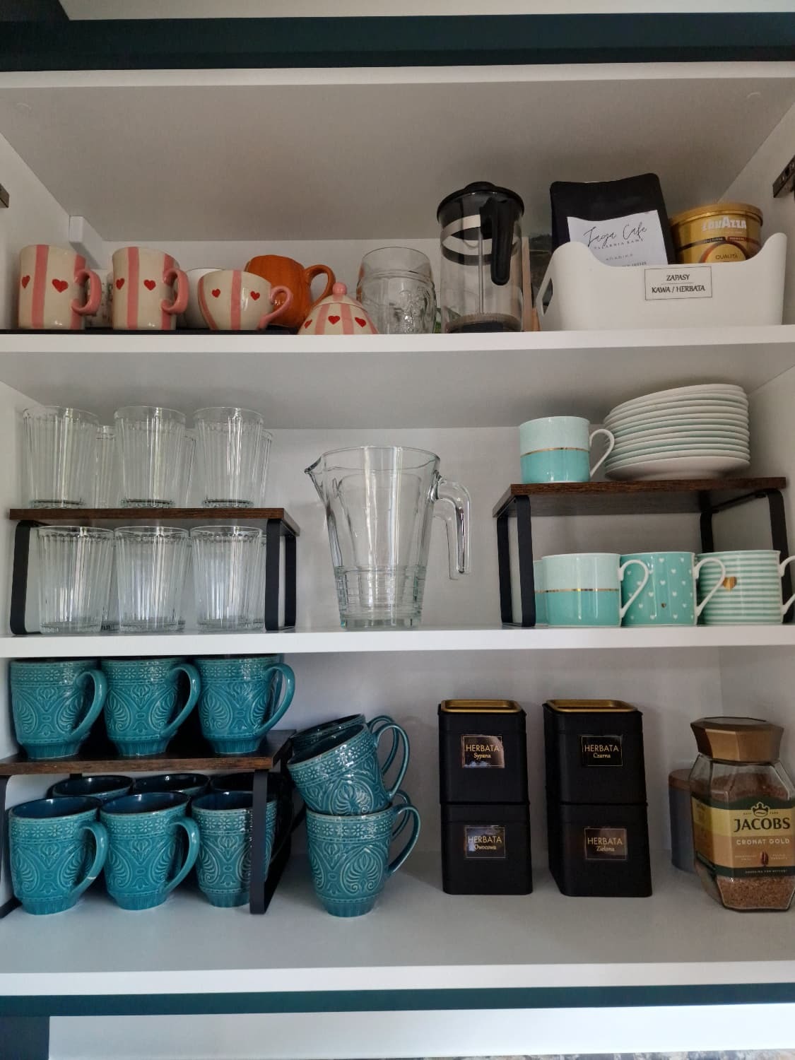 Uporządkowana szafka kuchenna z różnorodną zastawą: szklanki, dzbanki, filiżanki, puszki z herbatą i kawą. Estetyczne ułożenie naczyń na półkach.