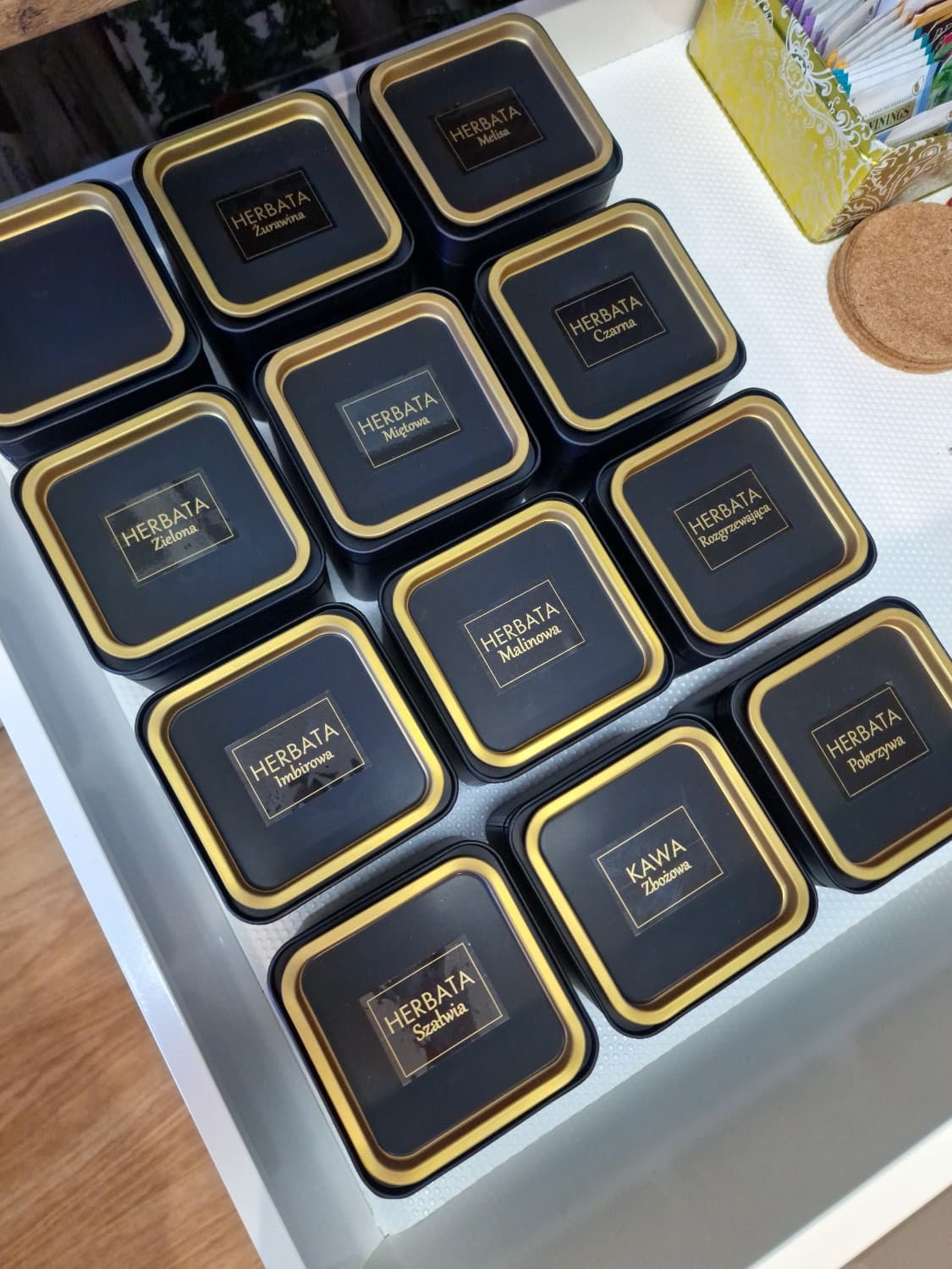 Kolekcja czarnych, kwadratowych puszek z herbatą i kawą zbożową, każda z etykietą w złotym obramowaniu, ułożonych w szufladzie. Różne smaki herbat.