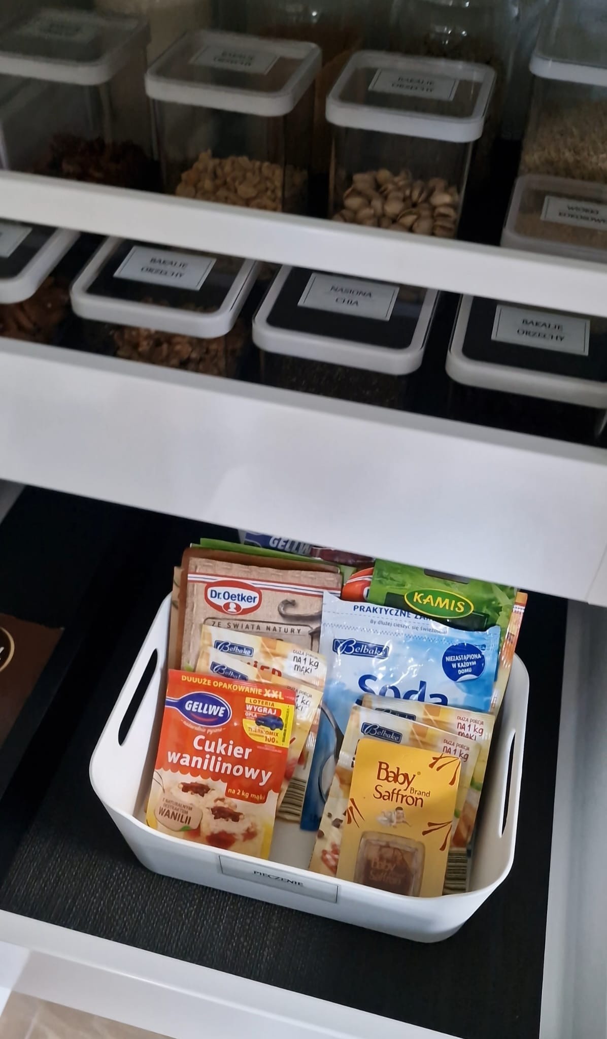 Uporządkowana szuflada z pojemnikami na bakalie i przyprawy w białym organizerze. Widoczne etykiety na pojemnikach i opakowaniach.
