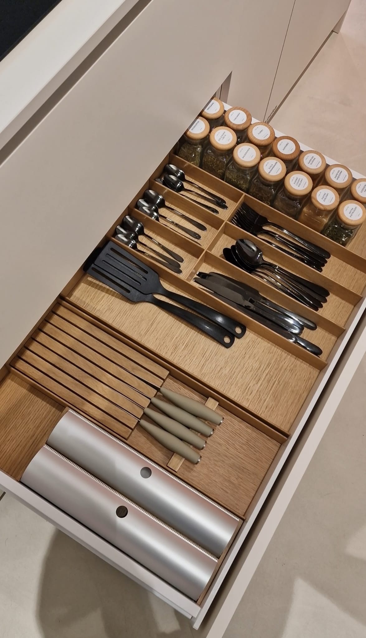 Otwarta szuflada kuchenna z drewnianym organizerem na sztućce, noże, folie aluminiowe i przyprawy w słoiczkach z etykietami. Porządek i organizacja w kuchni.