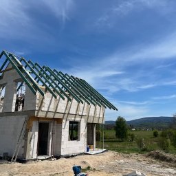 Piotr Kuczak - Wznoszenie dwukondygnacyjnego domu z białych bloczków, z widoczną konstrukcją dachu na tle błękitnego nieba i zielonych wzgórz. Etap budowy w toku, widoczne rusztowania.