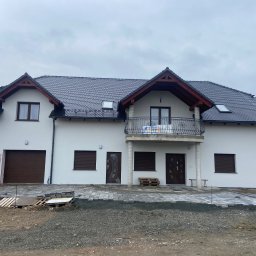 Piotr Kuczak - Nowoczesny, dwukondygnacyjny dom z balkonem i garażem w trakcie budowy. Częściowo ułożona kostka brukowa przed budynkiem. Szara dachówka i biała elewacja.