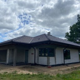 Piotr Kuczak - Niedokończony dom jednorodzinny z szarej cegły, z ciemną dachówką i oknami, w otoczeniu zieleni i z pochmurnym niebem w tle. Widoczny stan surowy zamknięty.