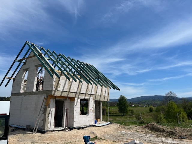 Wznoszenie dwukondygnacyjnego domu z białych bloczków, z widoczną konstrukcją dachu na tle błękitnego nieba i zielonych wzgórz. Etap budowy w toku, widoczne rusztowania.