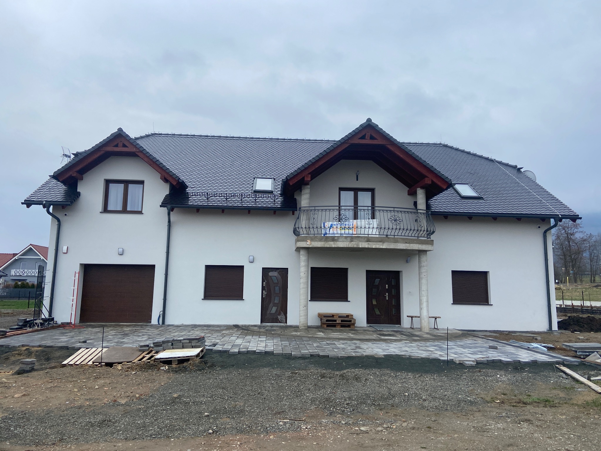 Nowoczesny, dwukondygnacyjny dom z balkonem i garażem w trakcie budowy. Częściowo ułożona kostka brukowa przed budynkiem. Szara dachówka i biała elewacja.