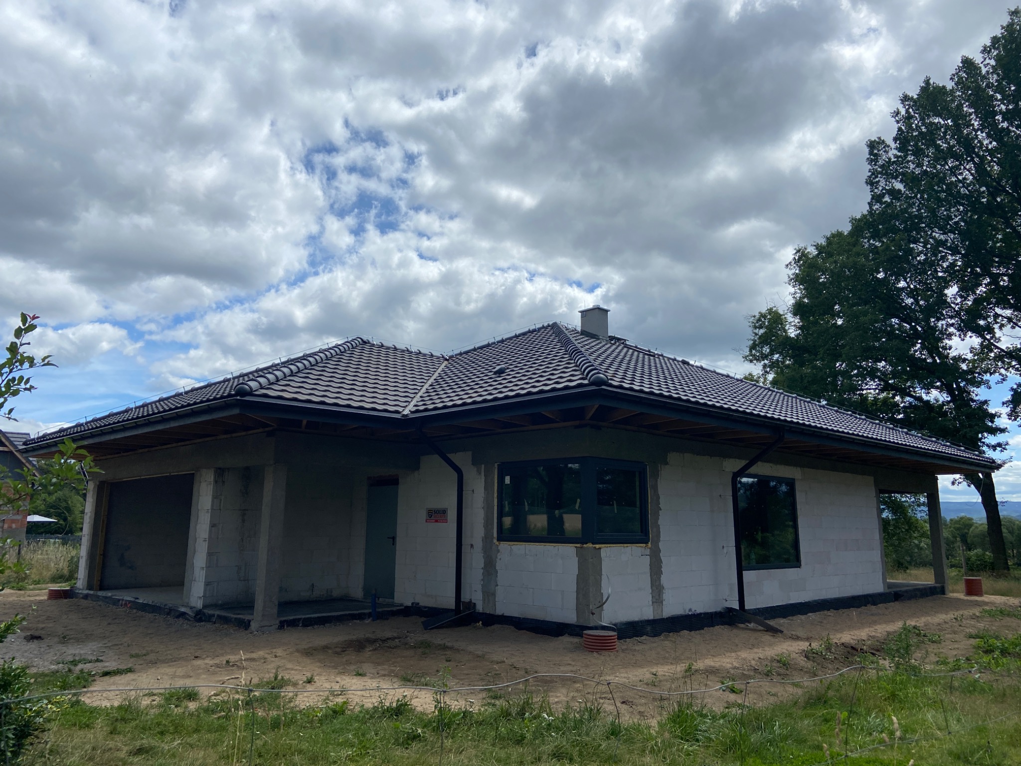 Niedokończony dom jednorodzinny z szarej cegły, z ciemną dachówką i oknami, w otoczeniu zieleni i z pochmurnym niebem w tle. Widoczny stan surowy zamknięty.