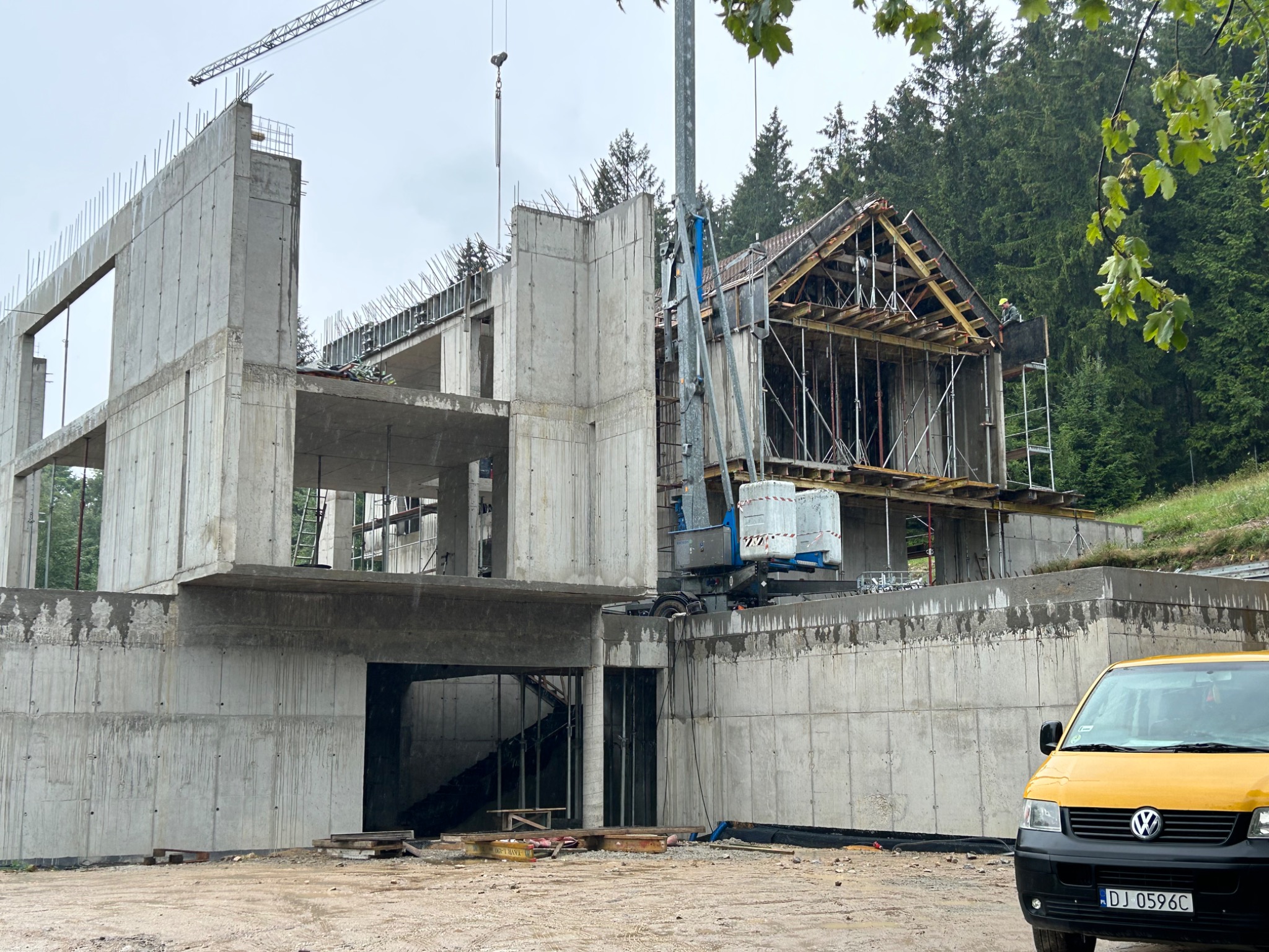 Surowy stan budynku żelbetowego w trakcie budowy, widoczny dźwig i szalunki, żółty samochód dostawczy na pierwszym planie, las w tle.
