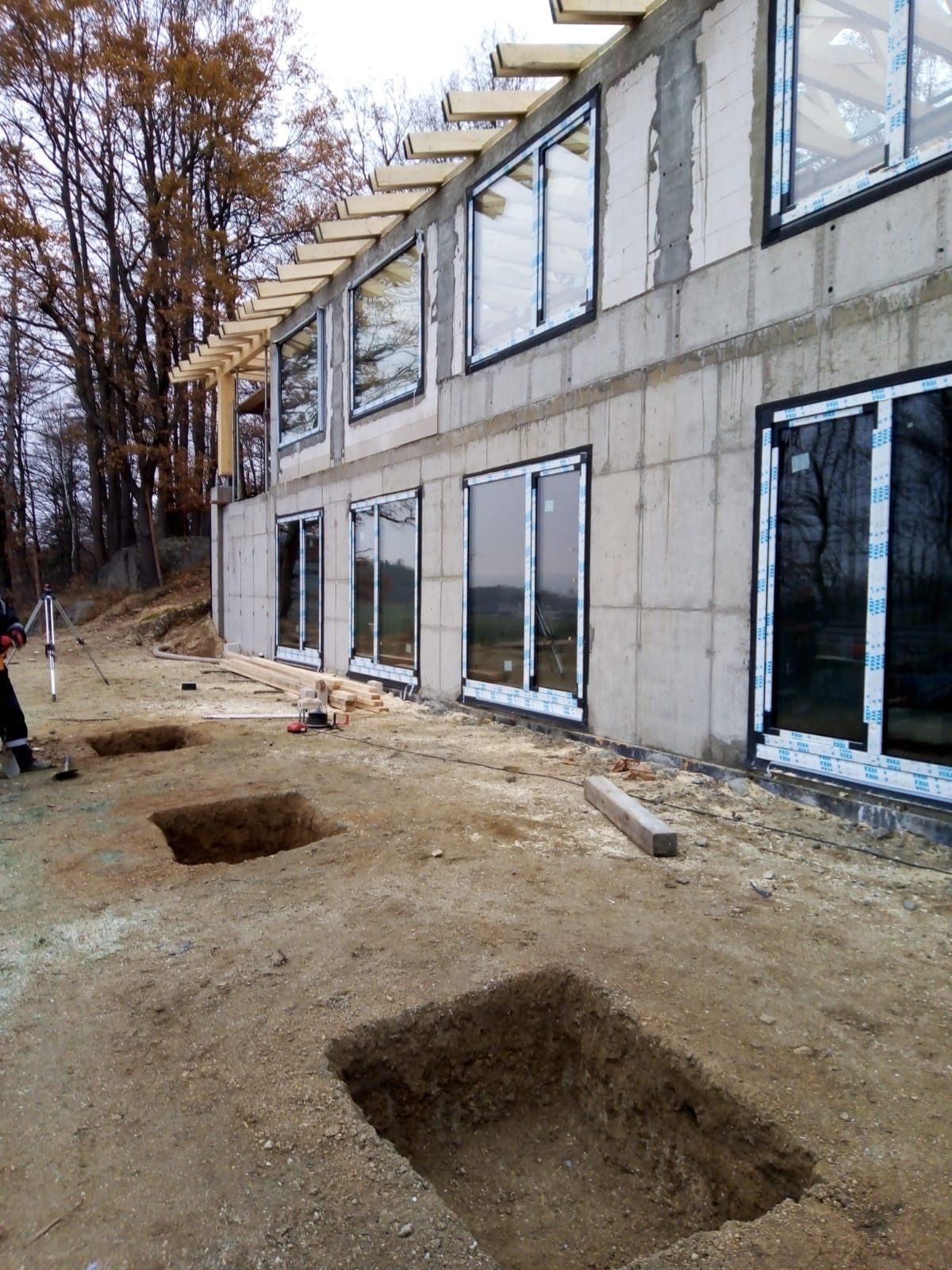 Surowy stan budynku z oknami i wykopami pod fundamenty. Widoczne drewniane elementy konstrukcji dachu. Prace ziemne w toku, jesienna sceneria w tle.