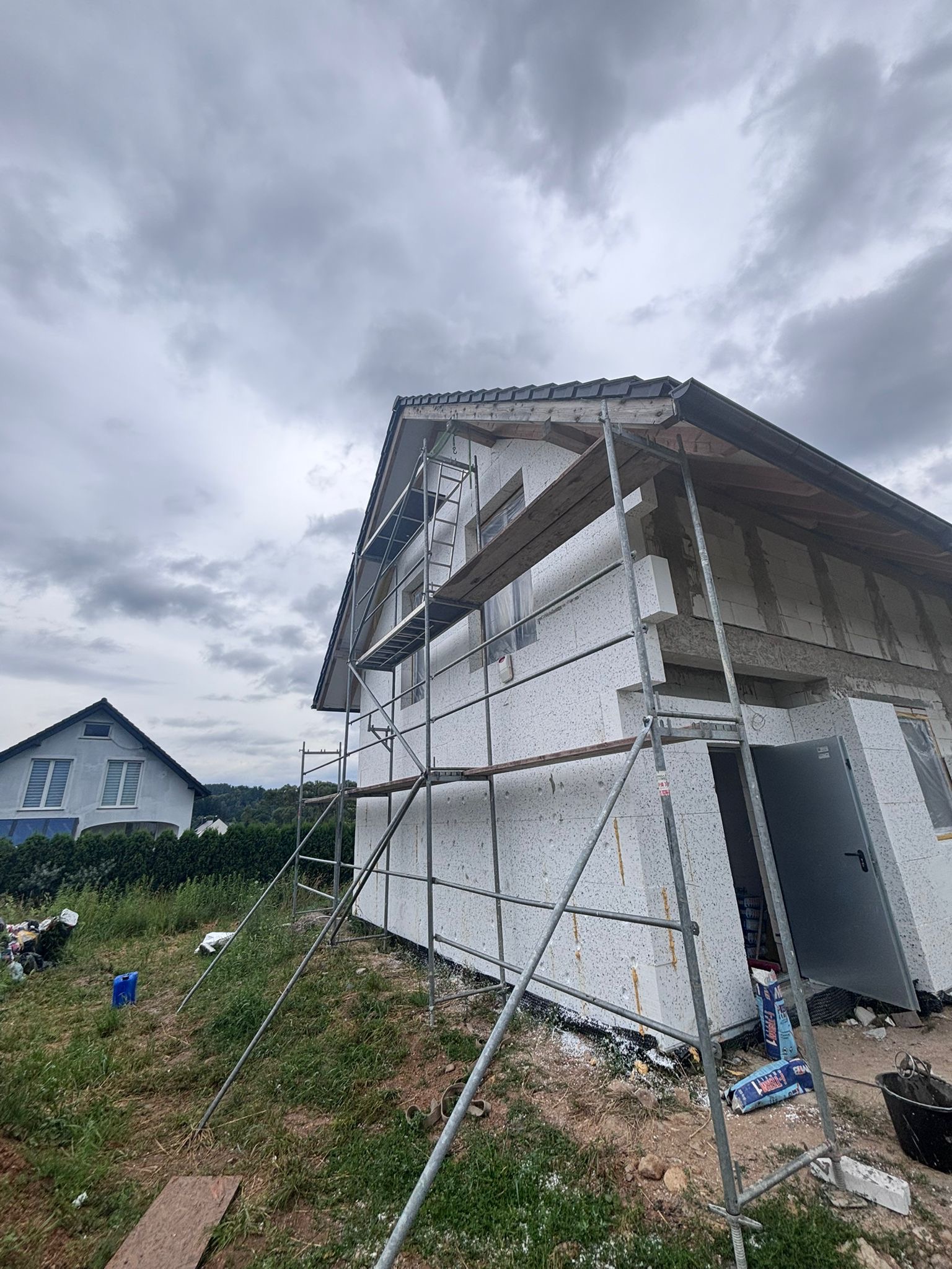 Dom w trakcie ocieplania, widoczne rusztowania i materiały izolacyjne. W tle drugi dom i pochmurne niebo. Ujęcie z dołu, podkreślające wysokość budynku.