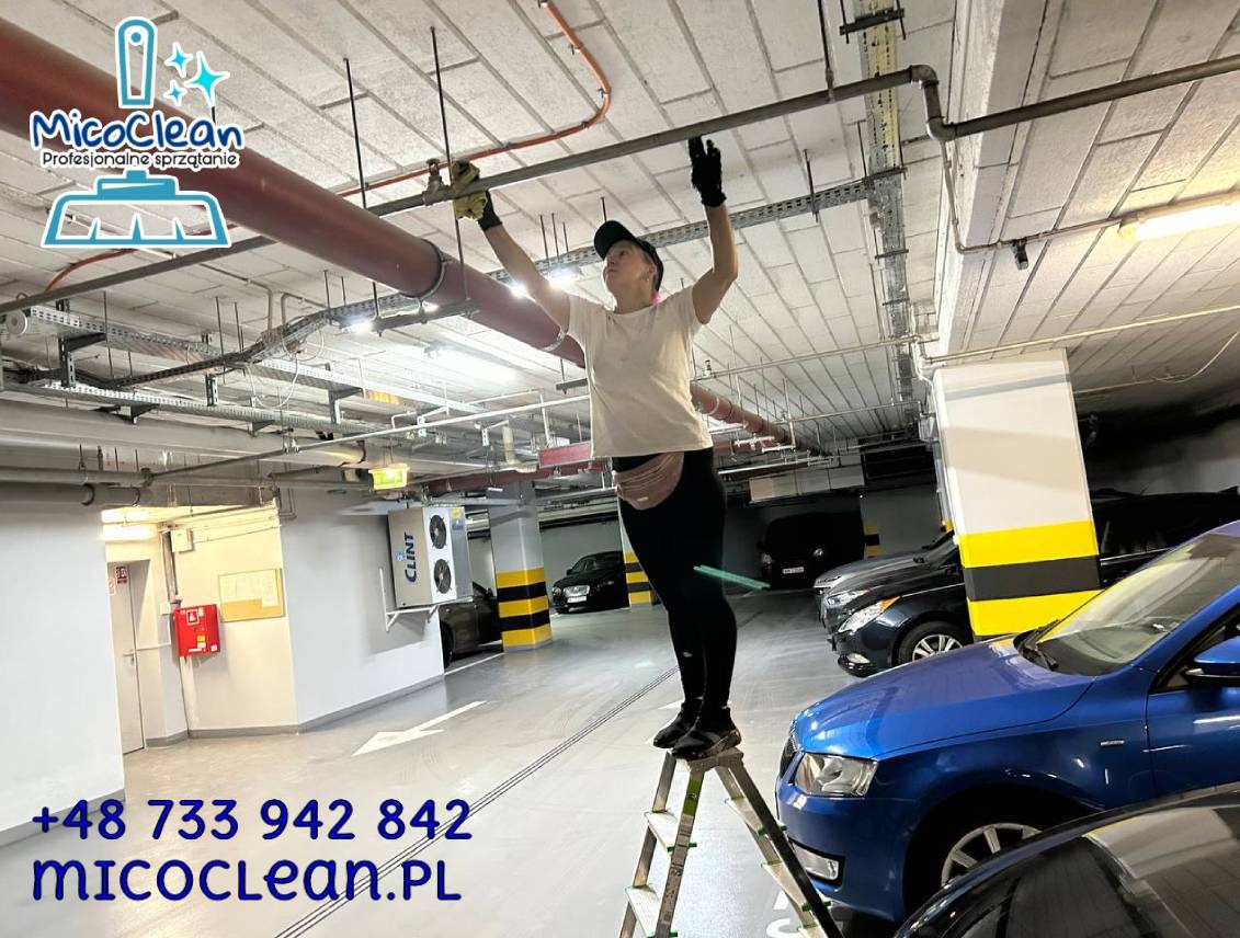 Kobieta na drabinie czyści rury pod sufitem parkingu podziemnego. Widoczne logo firmy sprzątającej i dane kontaktowe. Nietypowe ujęcie z dołu.
