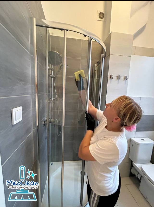 Osoba w czarnych rękawicach czyści kabinę prysznicową żółtą gąbką w łazience z szarymi płytkami. Widoczny fragment toalety i logo firmy sprzątającej.