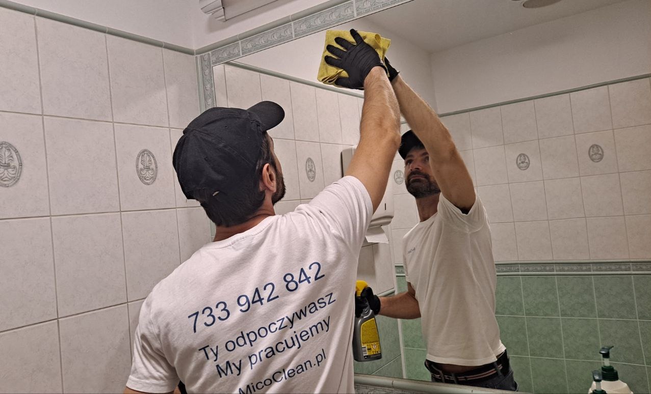 Pracownik w czarnej czapce i koszulce z numerem telefonu czyści lustro w łazience żółtą ściereczką, trzymając w dłoni butelkę ze środkiem czyszczącym.
