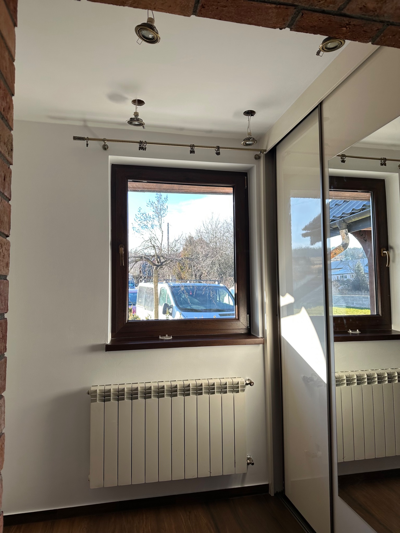 Jasne wnętrze po remoncie: białe ściany, okno z widokiem na zewnątrz, grzejnik i szafa z lustrzanymi drzwiami. Widoczne punkty świetlne na suficie.