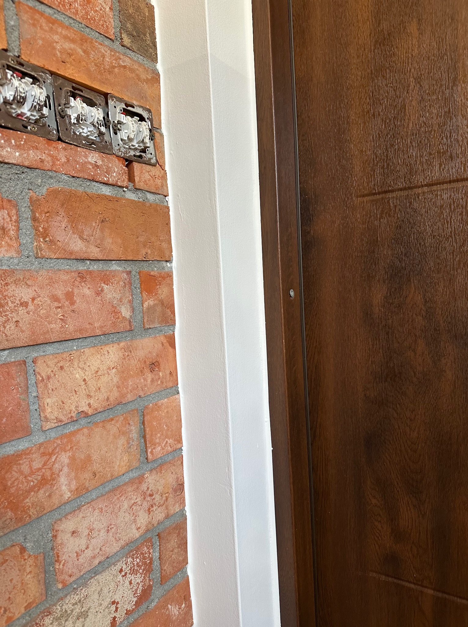 Fragment wnętrza: ceglana ściana z puszkami elektrycznymi, pomalowana na biało listwa przypodłogowa i drewniane drzwi. Detal wykończenia wnętrza.