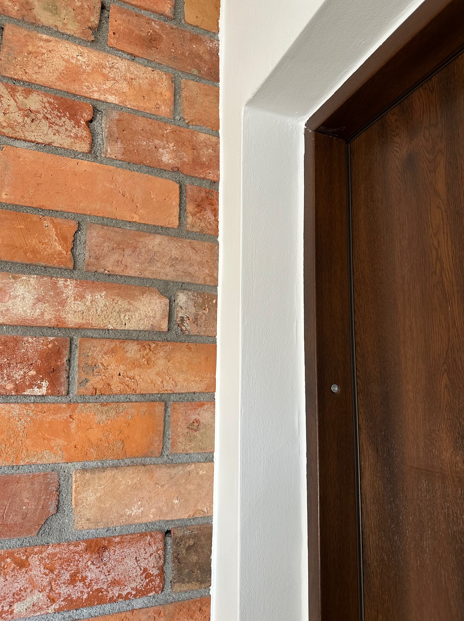 Wykończenie wnętrza: dekoracyjna ściana z cegły klinkierowej sąsiaduje z ościeżnicą pomalowaną na biało i drewnianymi drzwiami. Detal wykończeniowy.