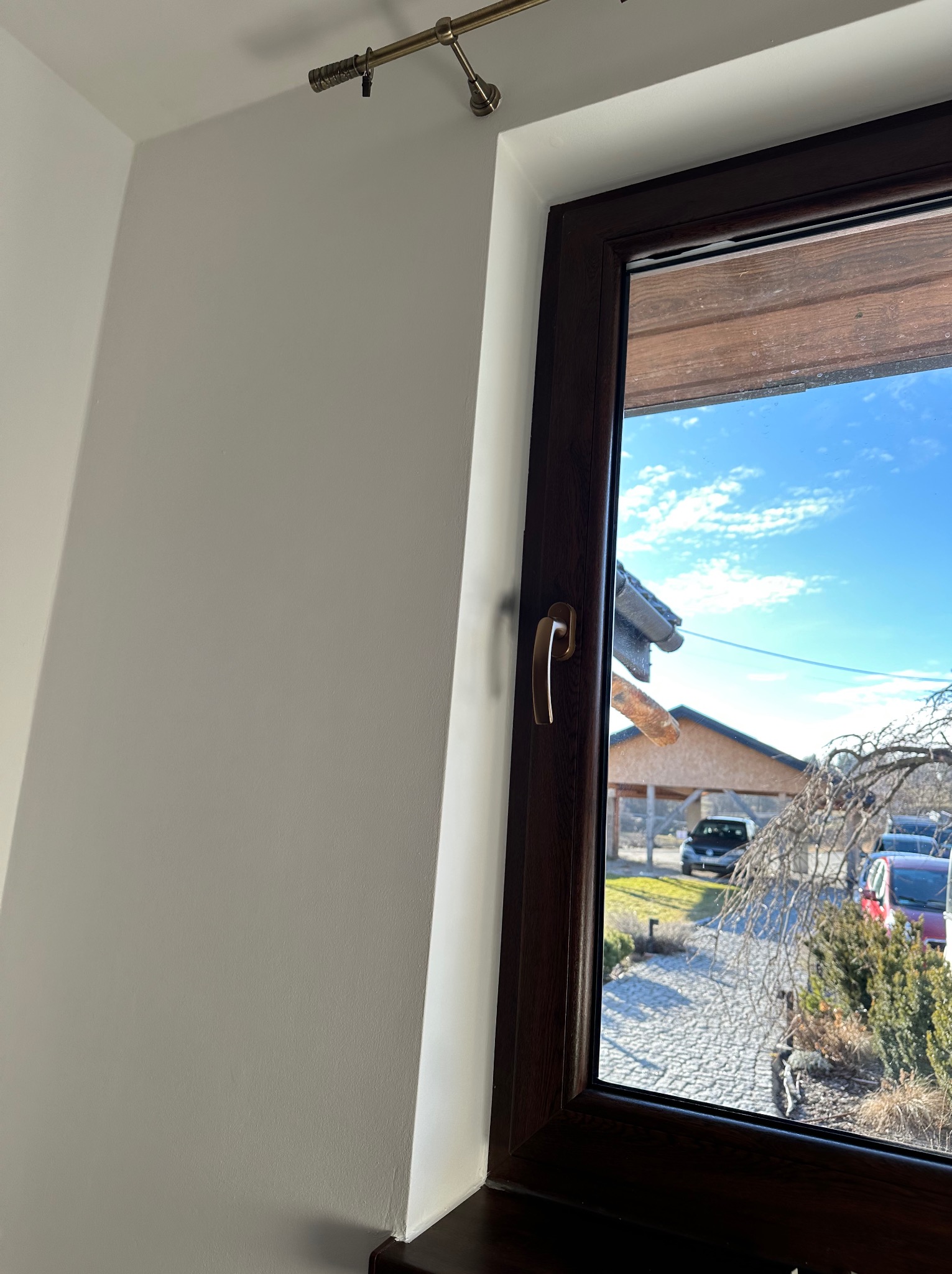 Świeżo pomalowana, gładka ściana w odcieniach bieli, widoczna z perspektywy okna z brązową ramą. Na ścianie widoczny karnisz. Widok na zewnątrz przez okno.