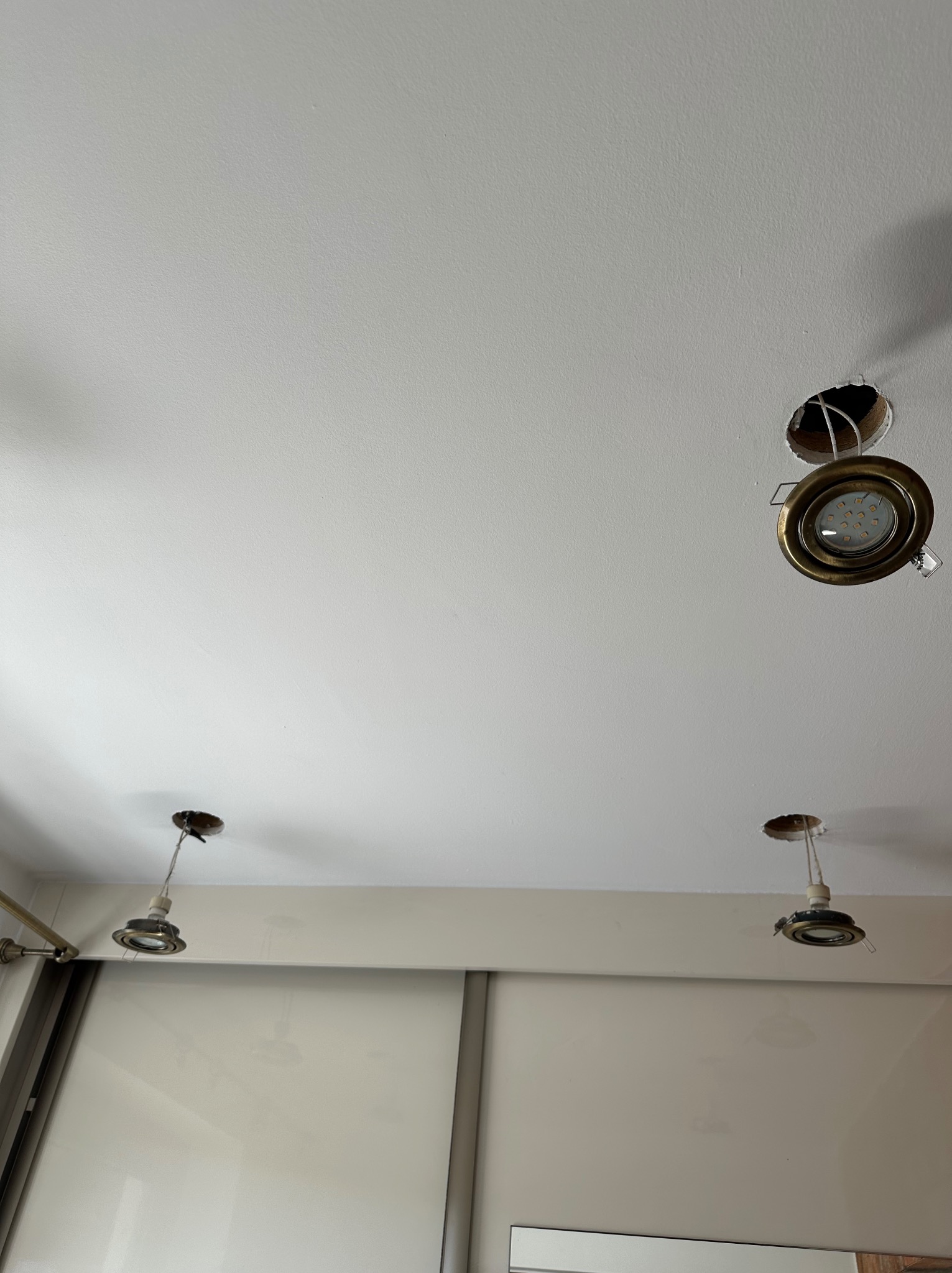 Biały sufit z otworami po halogenach i wystającymi kablami, przygotowany do montażu oświetlenia. Widoczne ślady po starych oprawach.