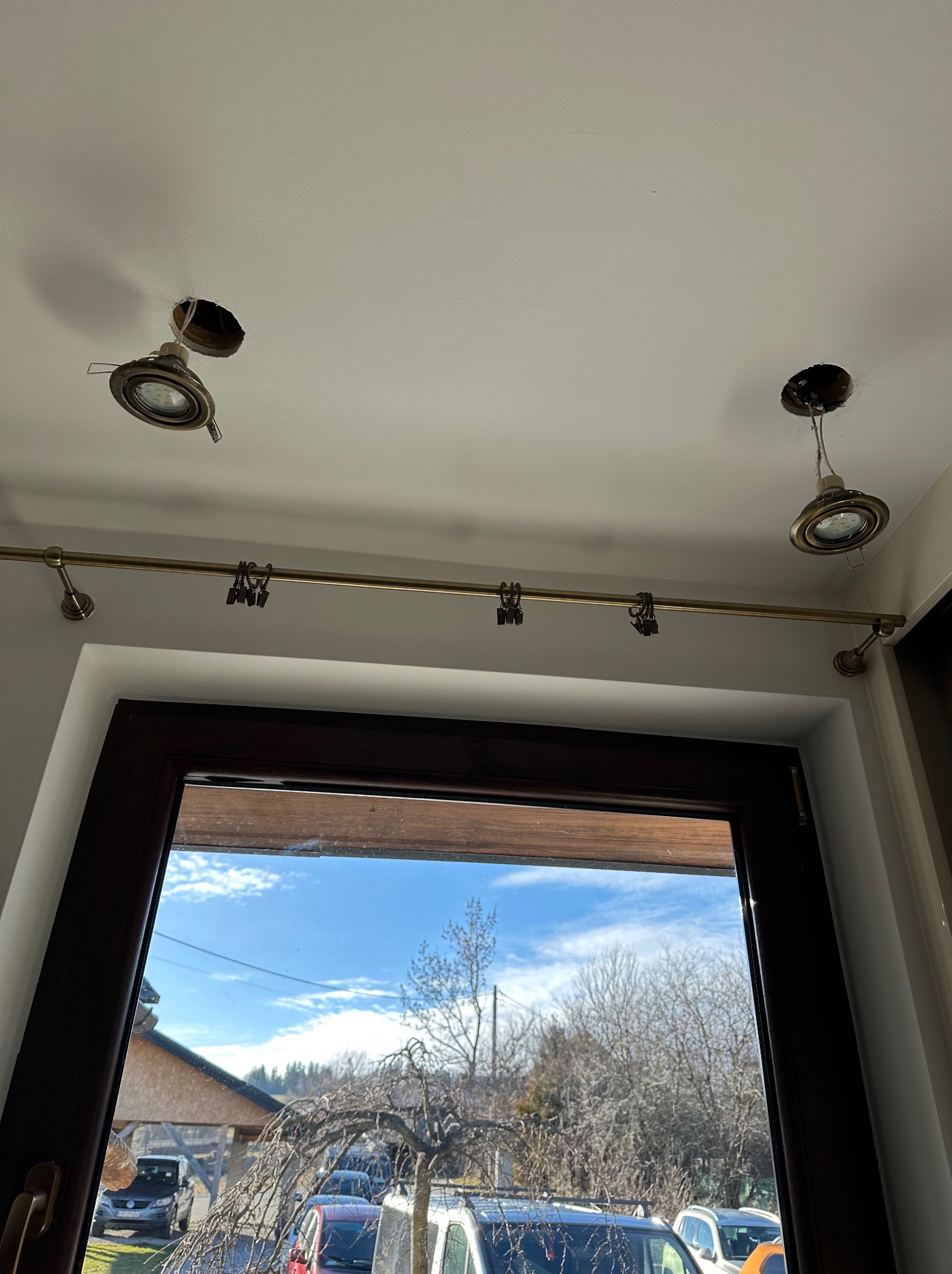 Montaż oświetlenia sufitowego w pokoju z oknem. Widoczne dwa punkty świetlne z odsłoniętymi przewodami i karnisz z klipsami. W tle widok na zewnątrz.