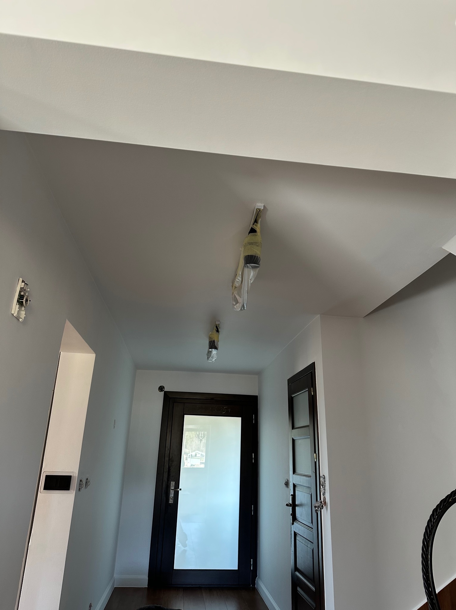 Jasne wnętrze po odświeżeniu: świeżo pomalowane ściany i sufit w odcieniach bieli, zabezpieczone lampy, widoczne drzwi i fragment schodów.