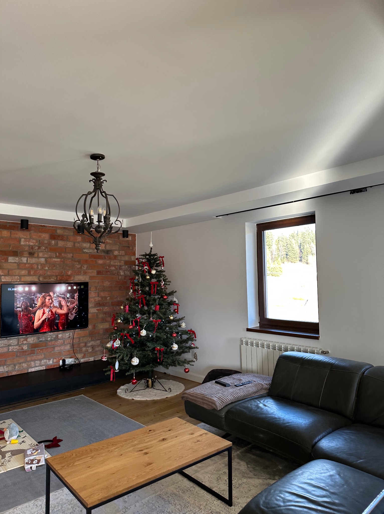 Salon z ceglaną ścianą, choinką i czarną sofą. Widoczne okno z widokiem na las. Na ścianie telewizor z włączonym programem. Całość w jasnych, naturalnych barwach.