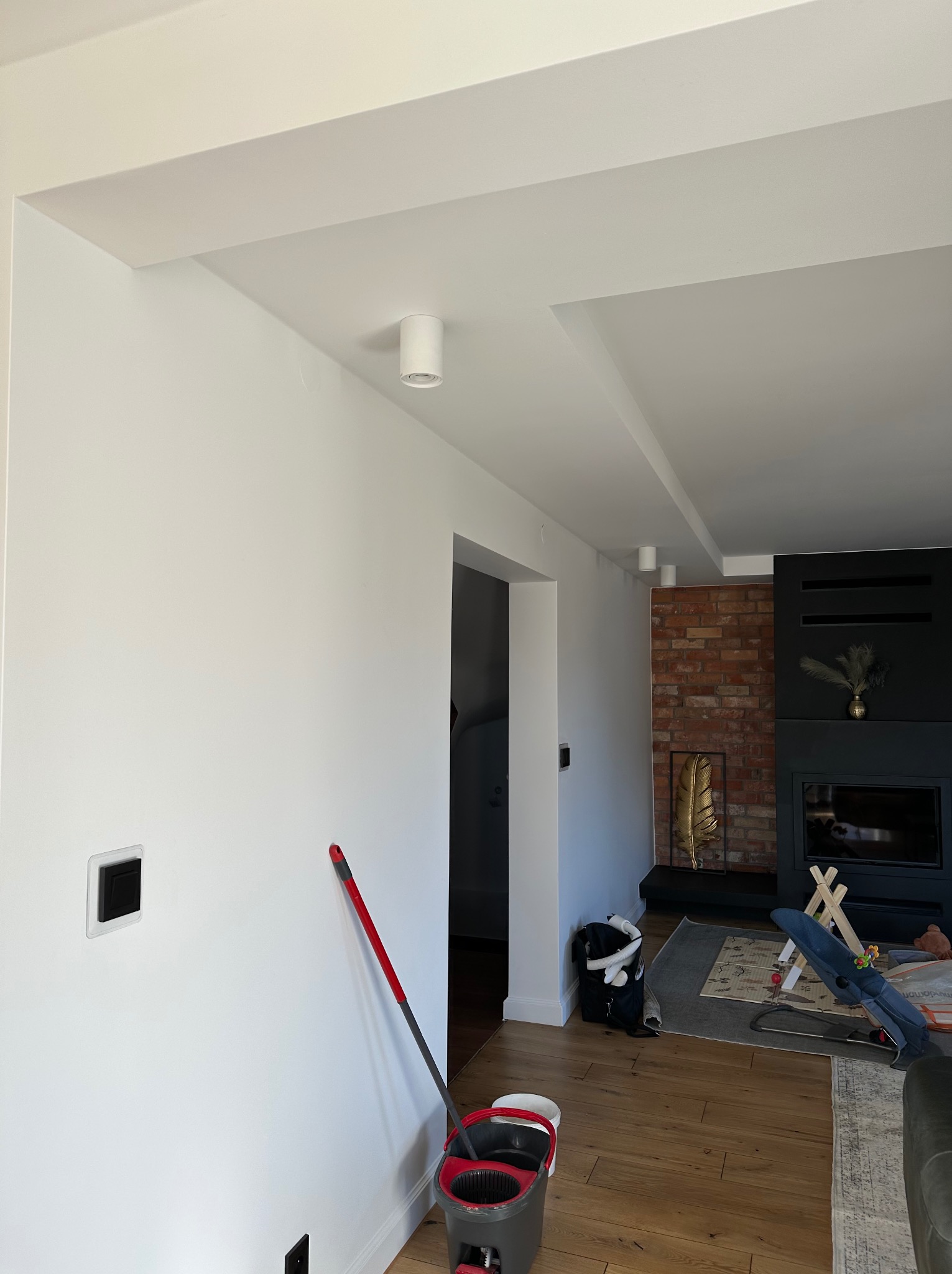 Białe ściany i sufit w salonie po remoncie, widoczny fragment podłogi, mop przy ścianie, ceglana ściana z kominkiem w tle. Minimalistyczny design.