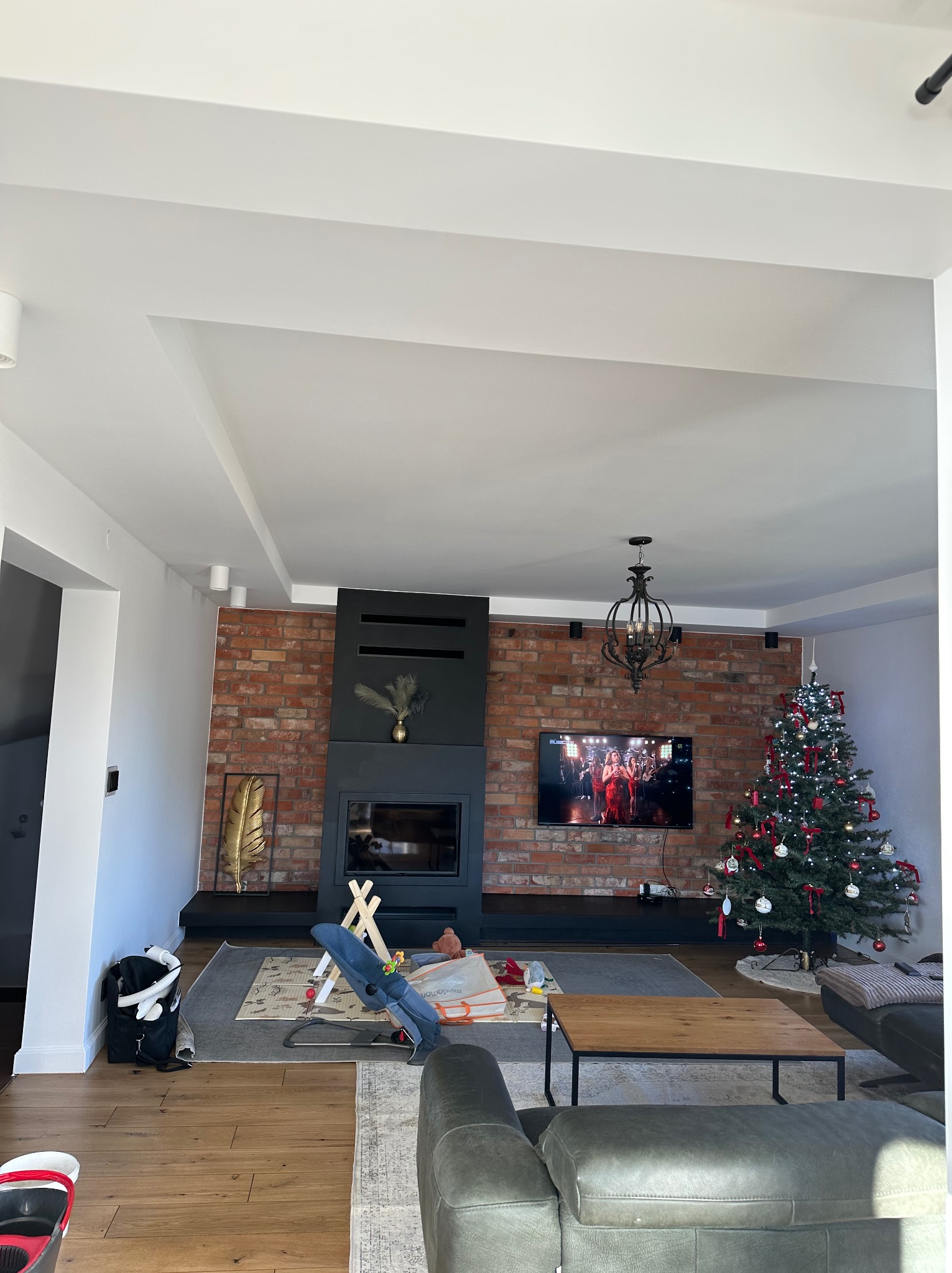 Salon z ceglaną ścianą, kominkiem i telewizorem. Ściany pomalowane na biało, sufit z podwieszanymi elementami. Widoczny fragment sofy i choinki.