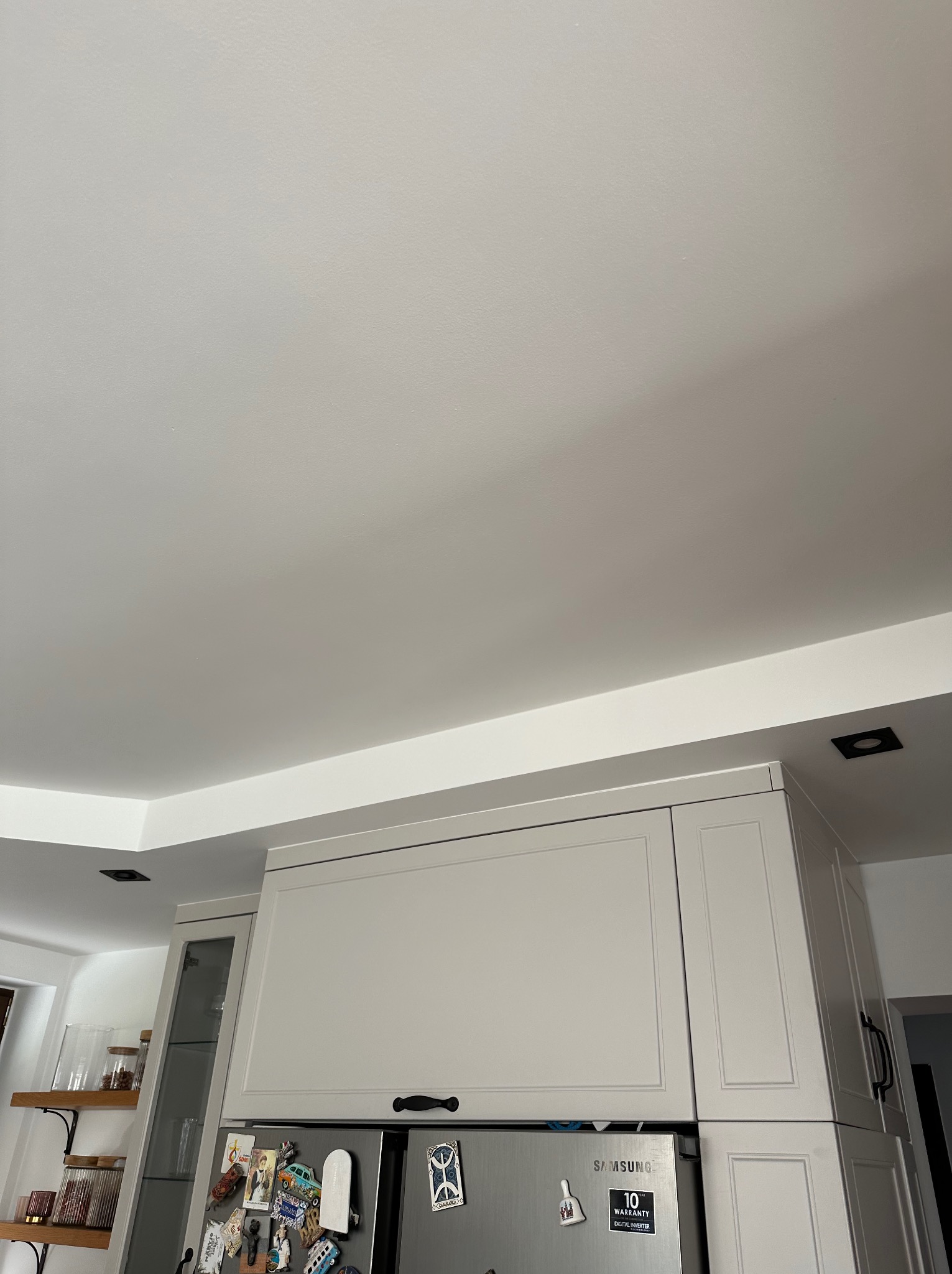 Gładka, biała powierzchnia sufitu po remoncie, z widocznymi elementami zabudowy kuchennej i oświetleniem punktowym. Minimalistyczny design wnętrza.