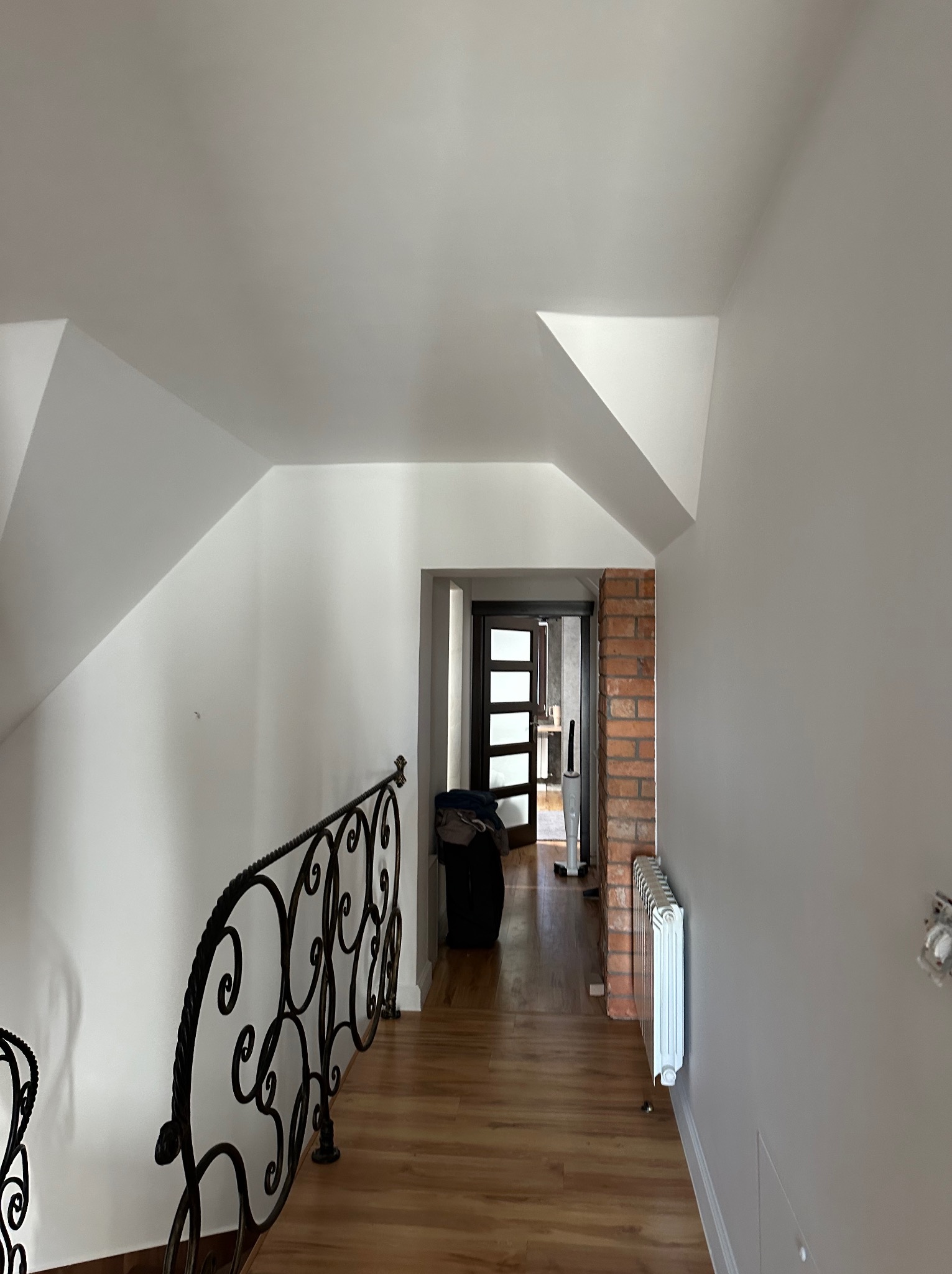 Białe ściany i sufit na poddaszu z ozdobną, kutą balustradą. Widok na korytarz z ceglanym wykończeniem i grzejnikiem. Świeżo pomalowane wnętrze.
