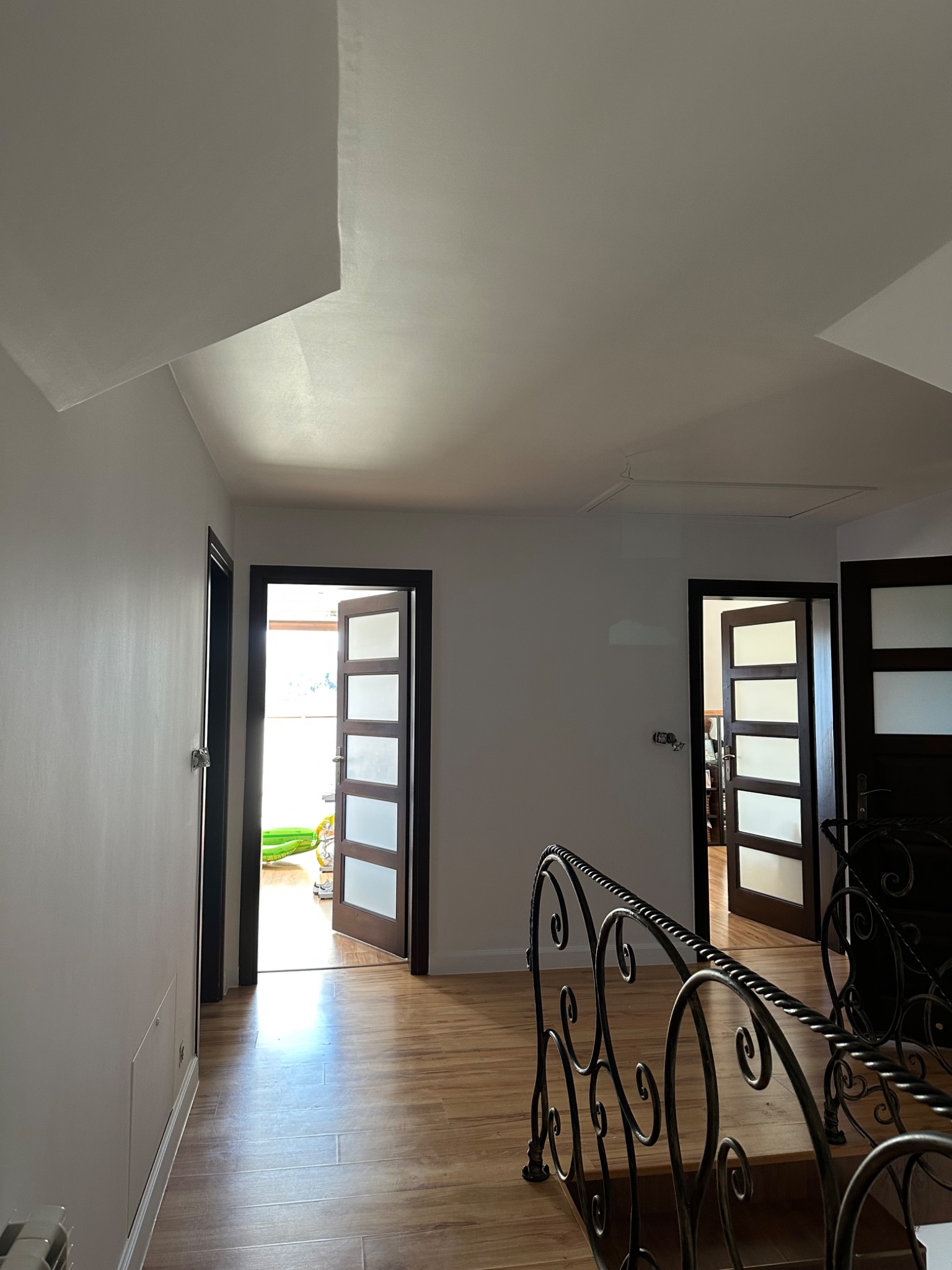 Jasne wnętrze domu po odświeżeniu: białe ściany, drewnopodobna podłoga, otwarte drzwi z przeszkleniami. Widoczne schody z metalową balustradą i fragment sufitu.