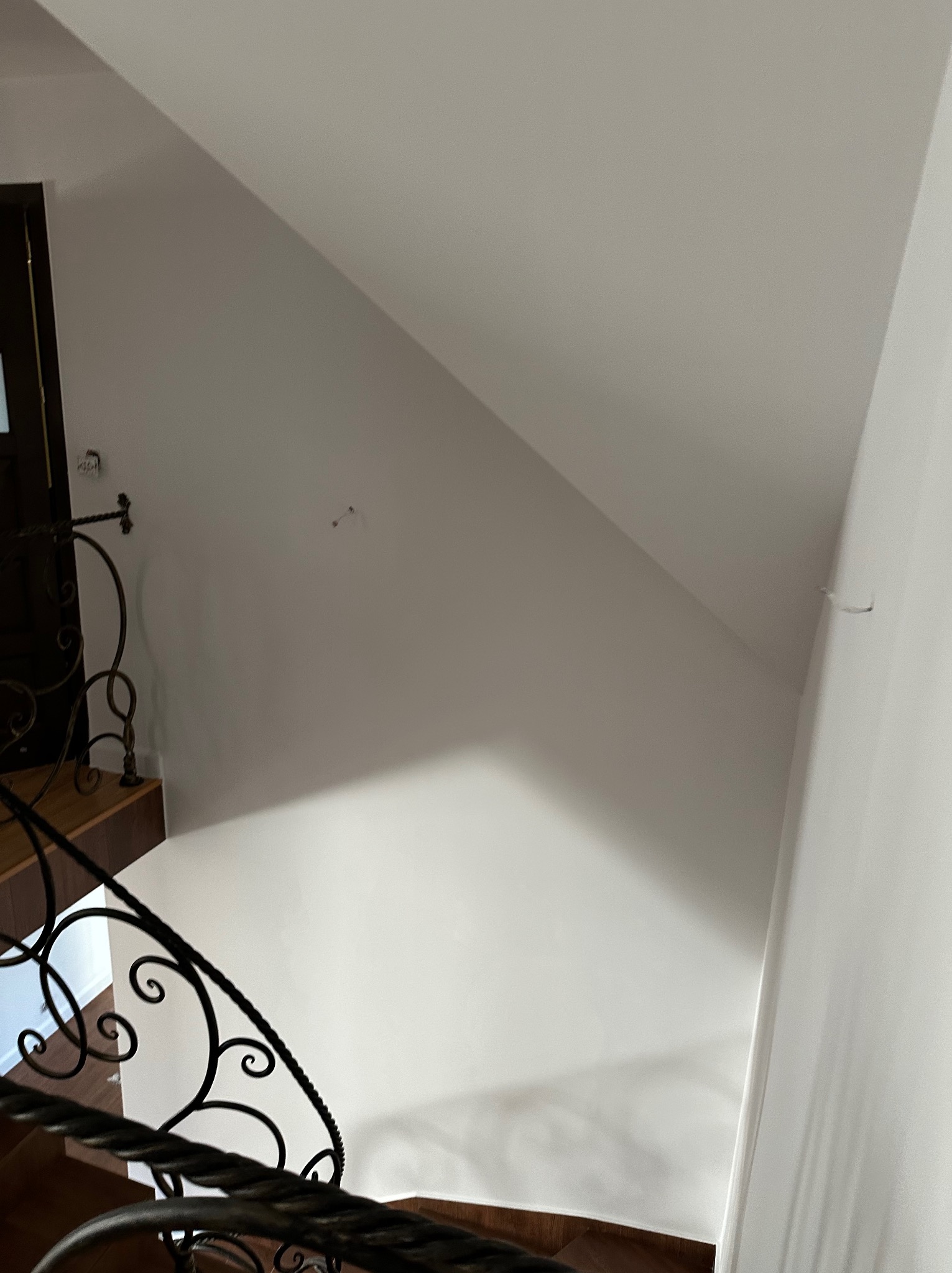 Biała ściana po malowaniu na poddaszu ze schodami z ozdobną, kutą balustradą. Widoczne gniazdko elektryczne i ślady po wkrętach.