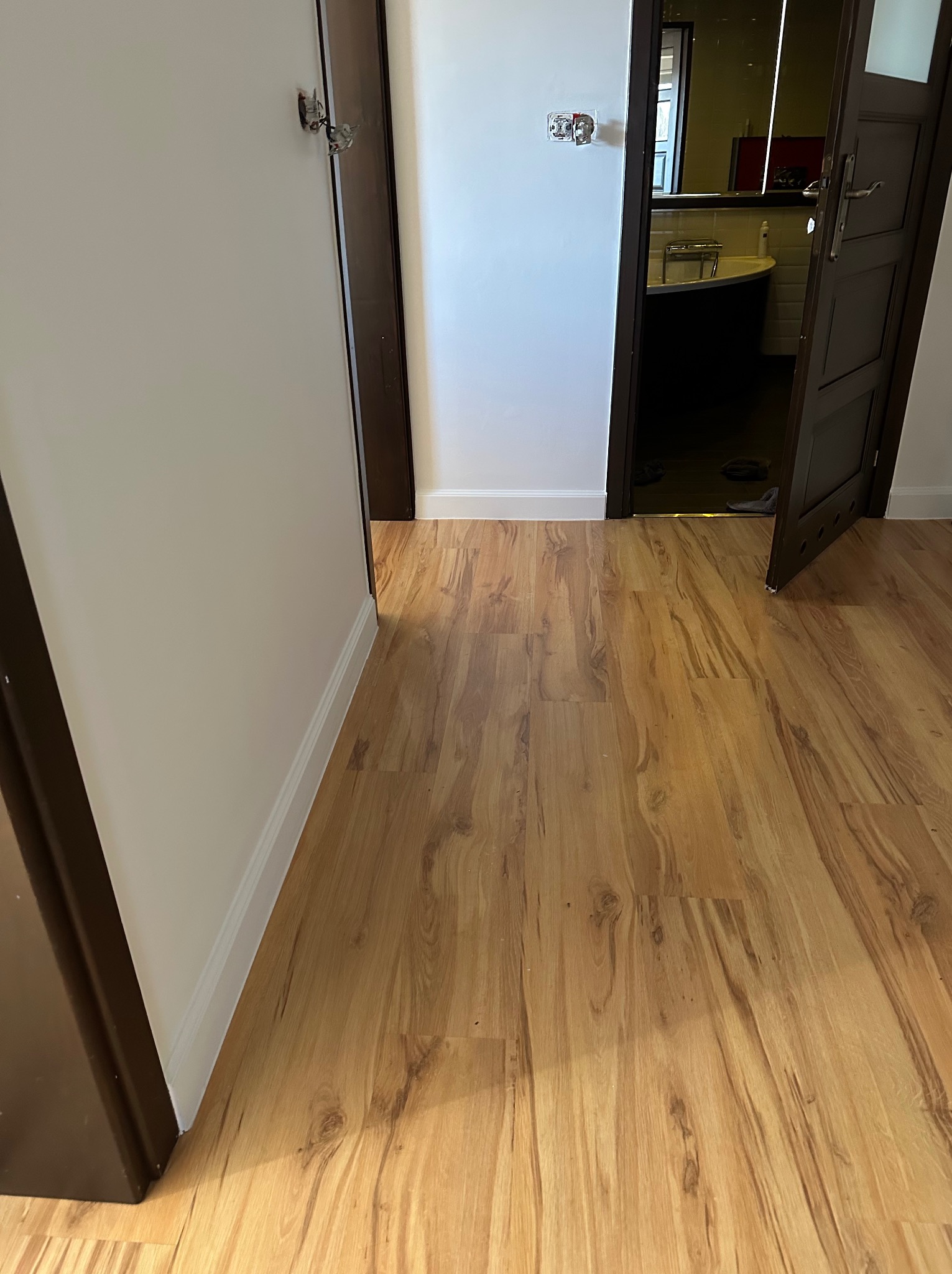 Jasne panele podłogowe w korytarzu z widokiem na łazienkę przez otwarte, ciemne drzwi. Ściany pomalowane na biało, widoczne listwy przypodłogowe.