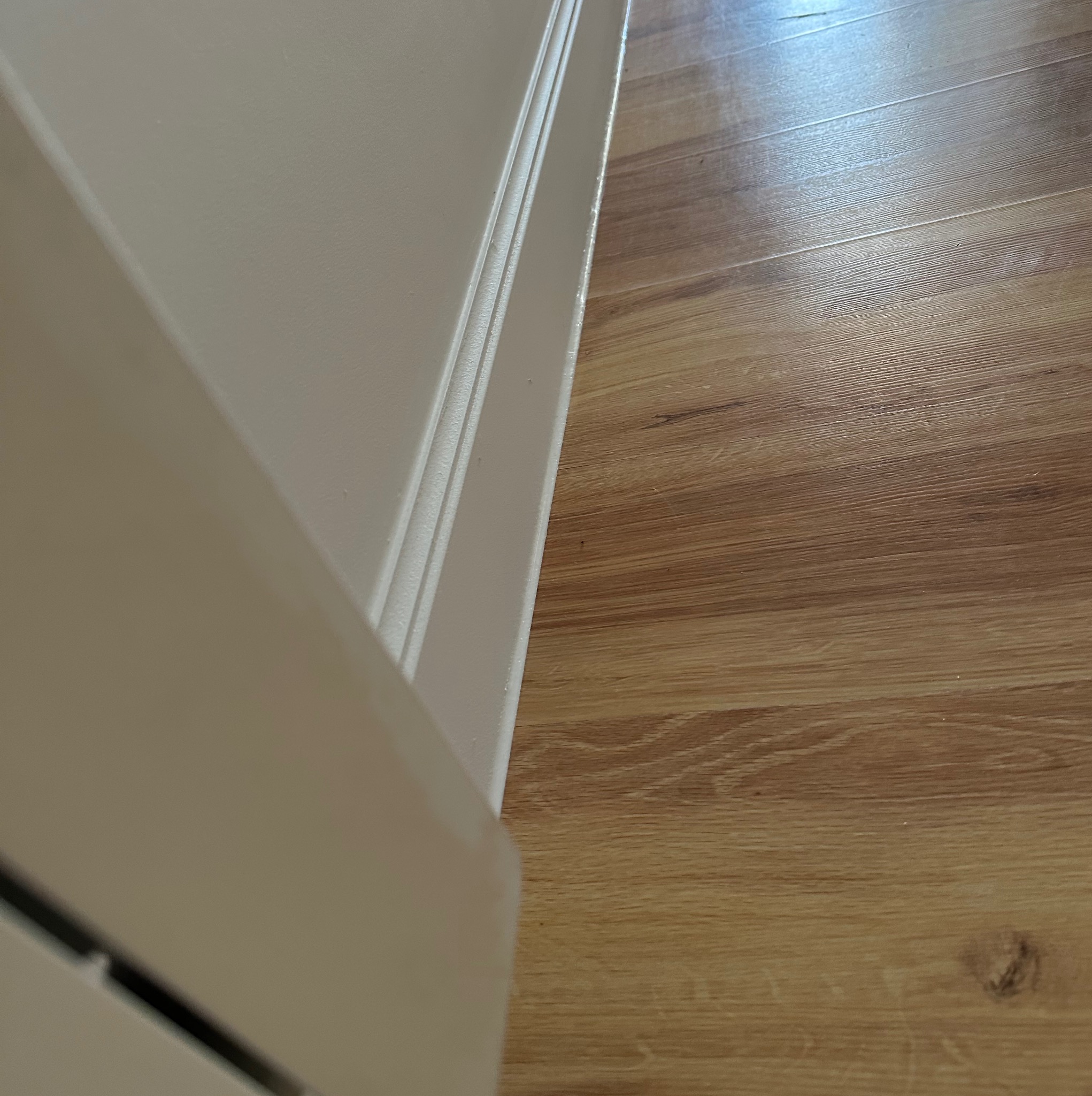 Świeżo pomalowana, kremowa ściana z listwą przypodłogową sąsiaduje z parkietem o ciepłym, drewnianym odcieniu. Widoczne detale wykończenia.