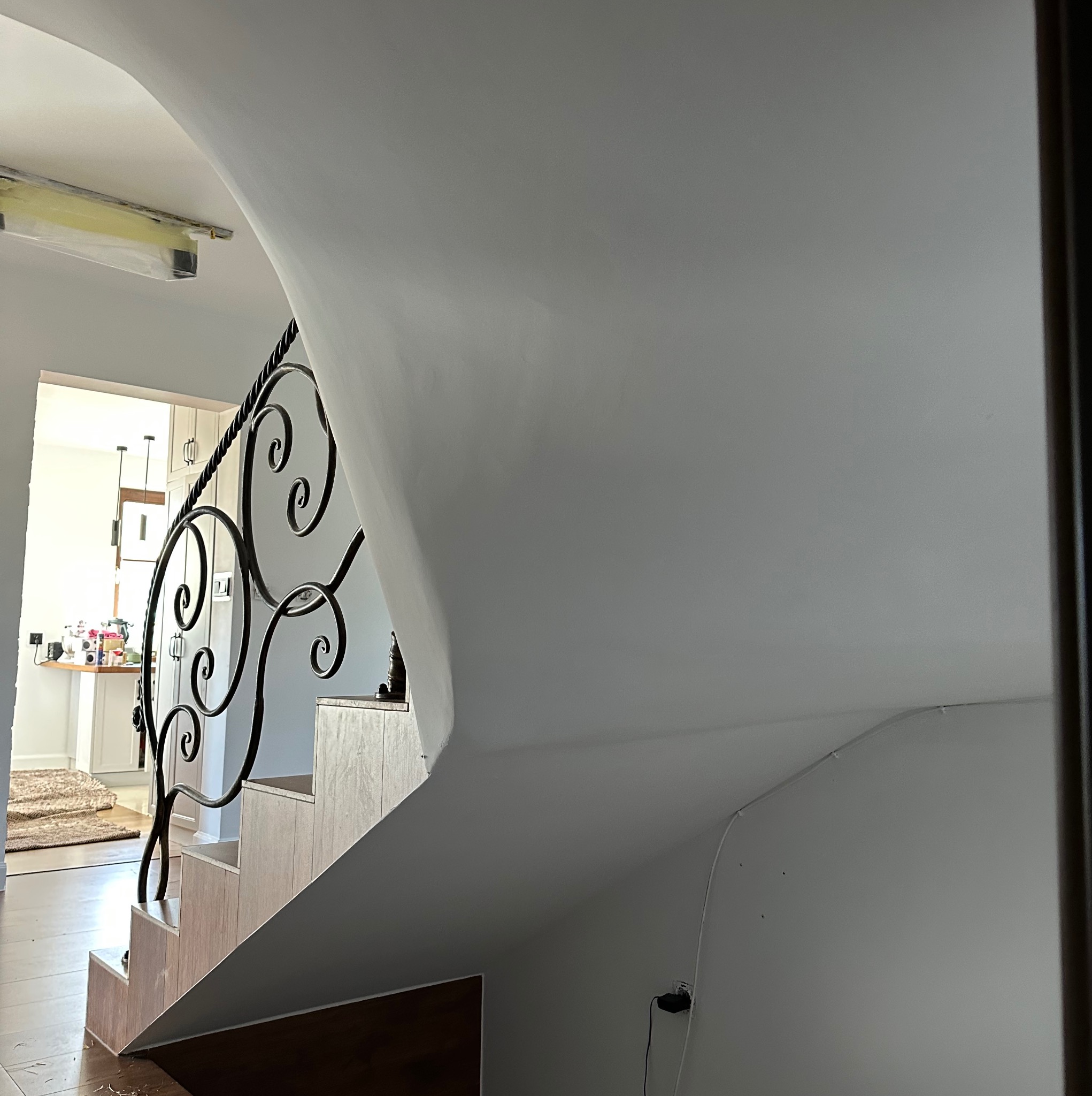 Gładka, biała ściana klatki schodowej z ozdobną, czarną balustradą. Widoczny fragment schodów i wnętrze domu w tle. Minimalistyczny design.