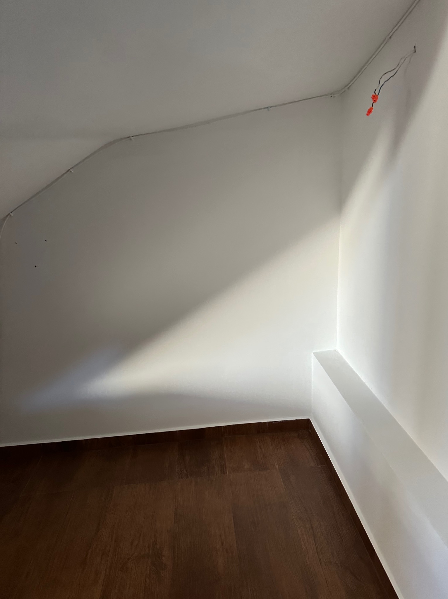 Wnętrze po remoncie: gładkie, białe ściany z widocznymi przewodami elektrycznymi, brązowa podłoga. Ukończone wykończenie wnęki z półką. Minimalistyczny design.