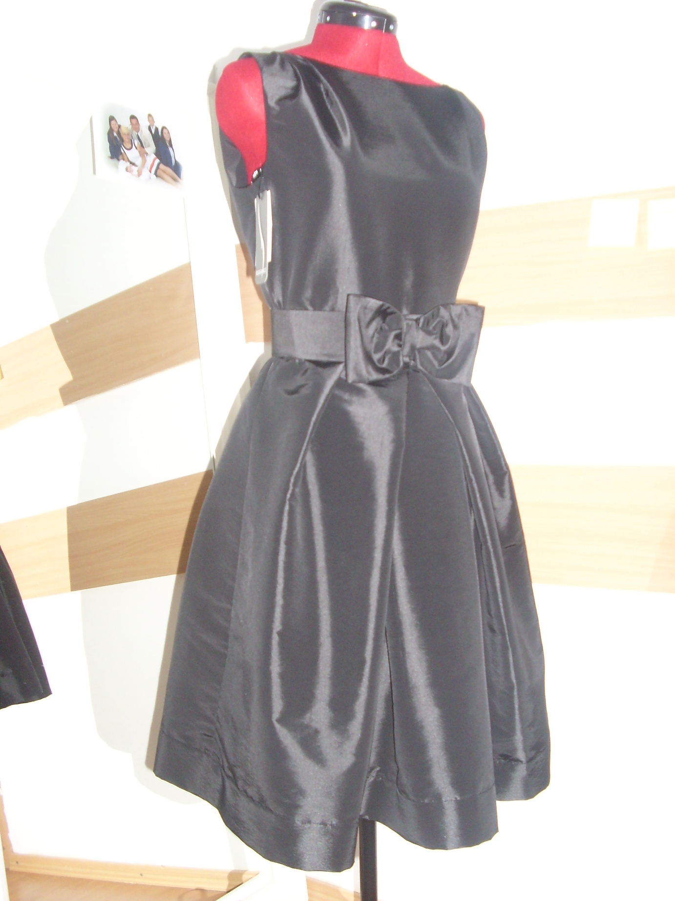 Elegancka, czarna sukienka z satyny z kokardą w pasie, prezentowana na czerwonym manekinie krawieckim, z widoczną fotografią grupy osób w tle.