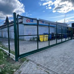 KC-SPAW - Zielone metalowe ogrodzenie z bramą przesuwną, zamontowane wzdłuż wybrukowanego podjazdu. W tle budynek hurtowni elektrycznej i samochód dostawczy.