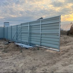 KC-SPAW - Szara brama przesuwna z blachy falistej na betonowej podstawie, w trakcie montażu na piaszczystym terenie, z traktorem i szklarnią w tle o zachodzie słońca.